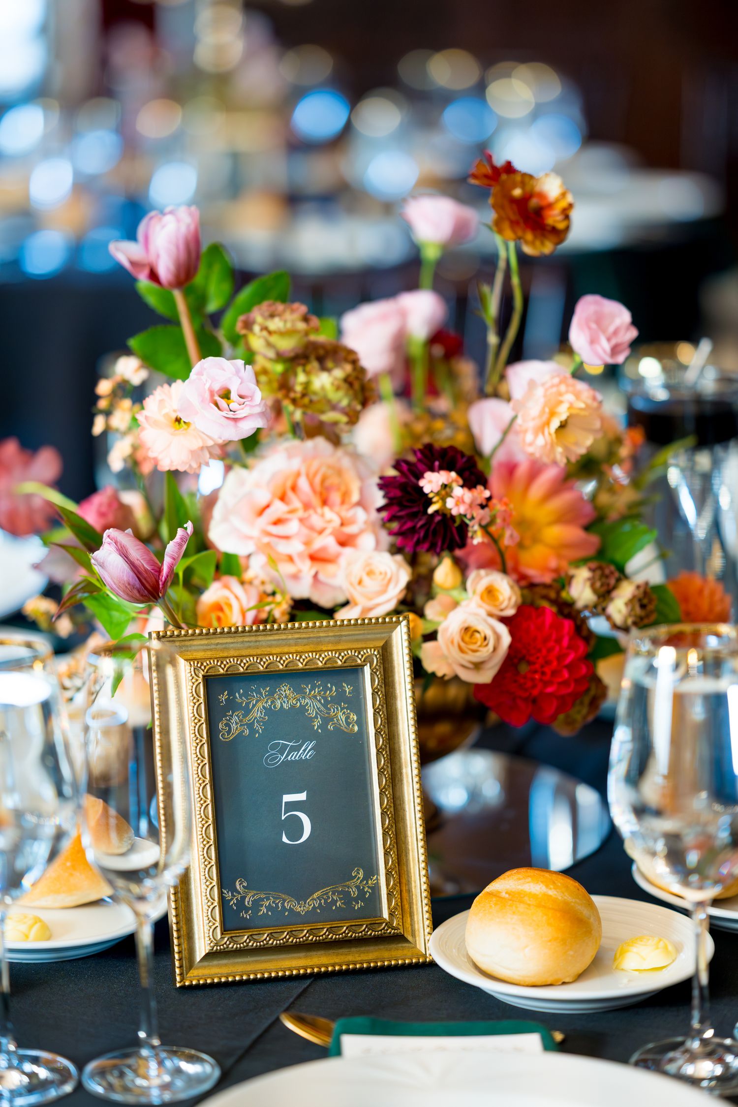 Elegant wedding reception table settings with gold-framed table numbers and coral floral centerpieces against dark tablecloths.