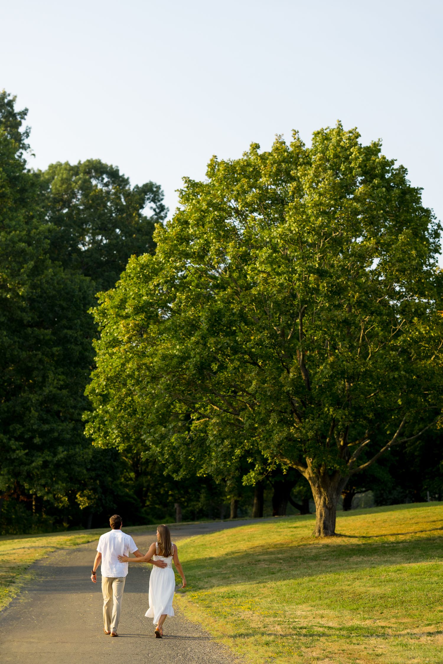 A couple walks hand in hand through a scenic park with mature trees on a sunny afternoon.