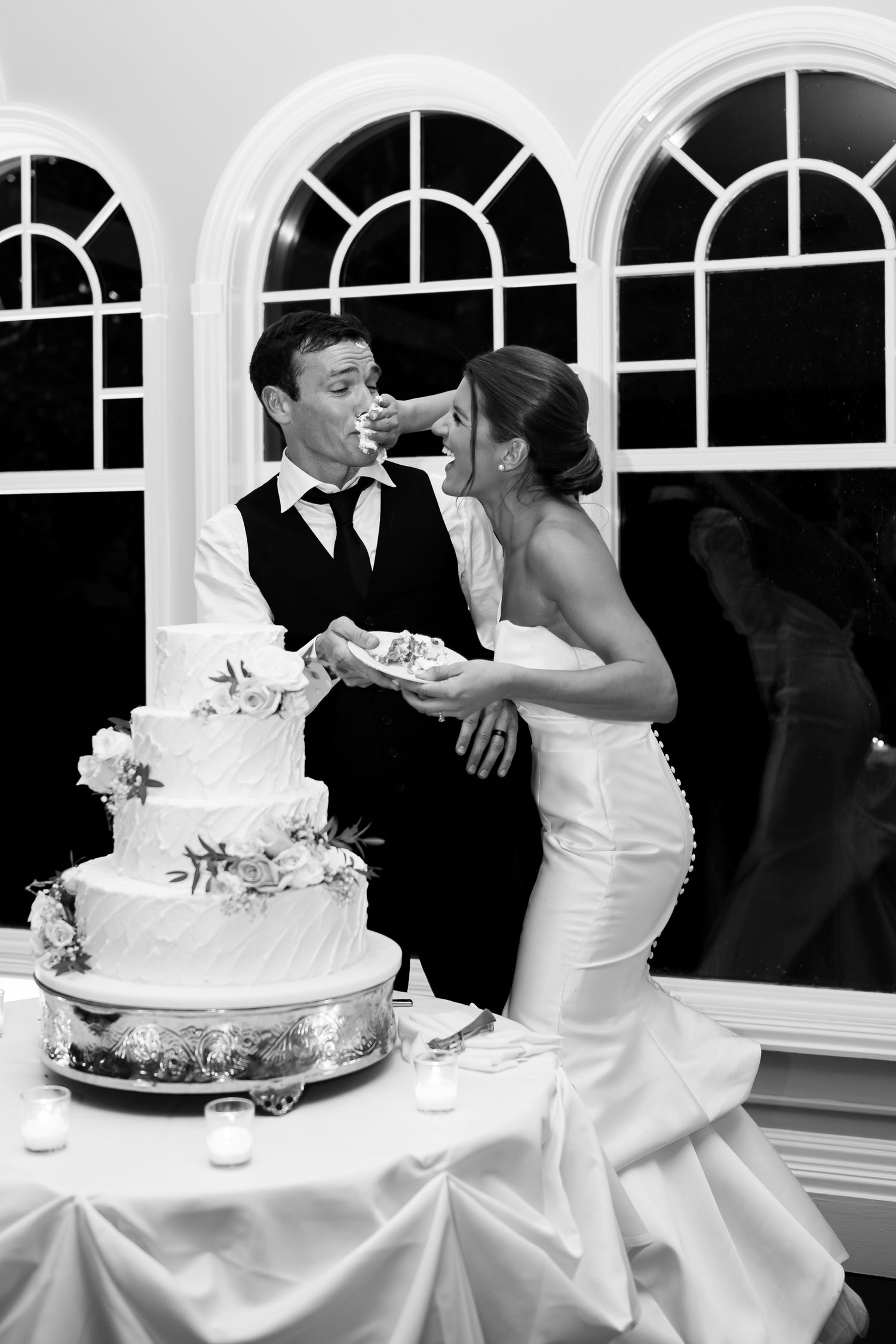 Black and white photo of couple sharing cake during wedding reception by arched windows.
