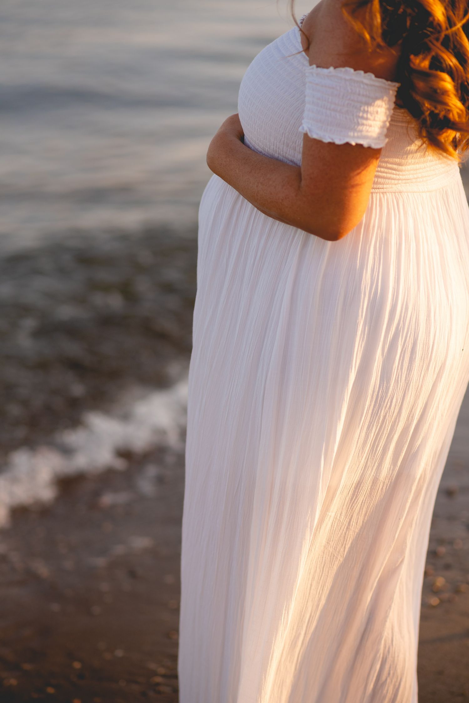 Women in flowing white dresses standing together on a beach at golden hour.