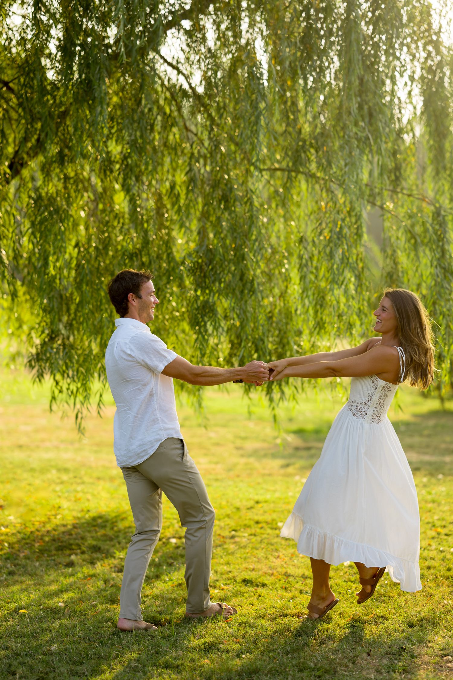 A couple playfully dances under a weeping willow tree at sunset, with the bride wearing a white dress.