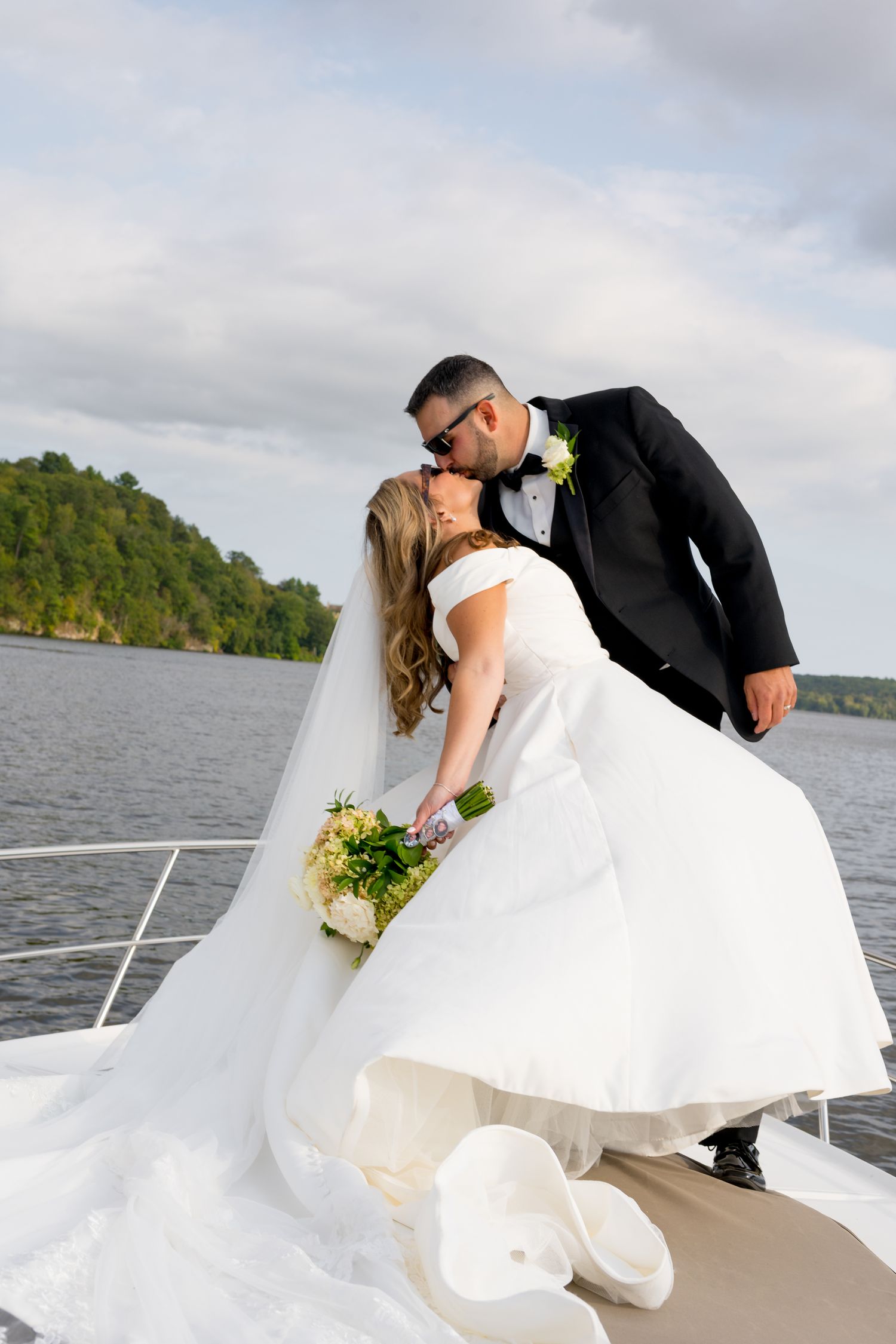 Romantic wedding couple shares intimate dip kiss moment on a boat deck with scenic lake and forest backdrop.