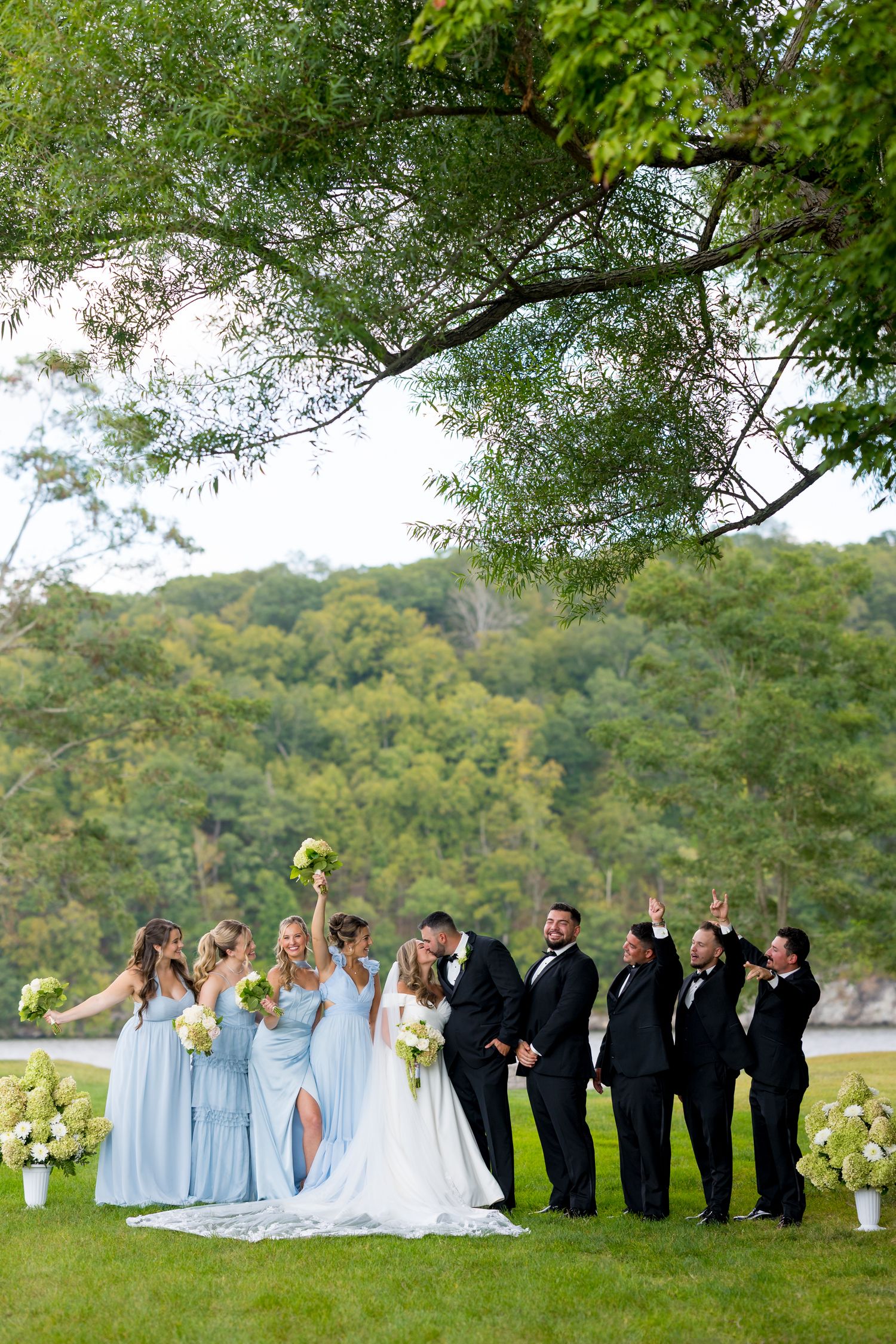 Wedding party in light blue dresses and black tuxedos poses on grassy hillside with trees in background.