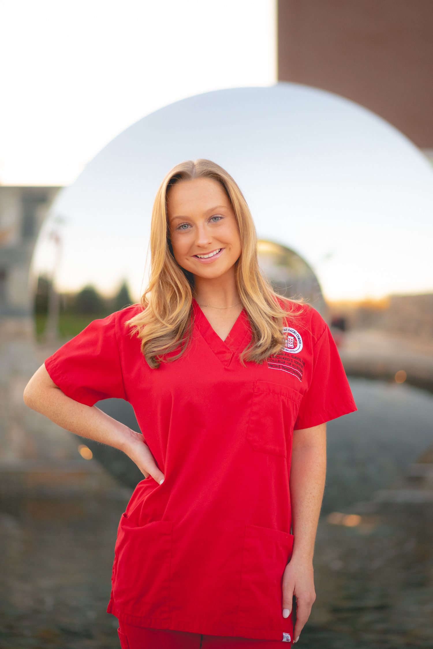 Sacred Heart nursing graduate in red scrubs poses outdoors during golden hour for graduation photos.