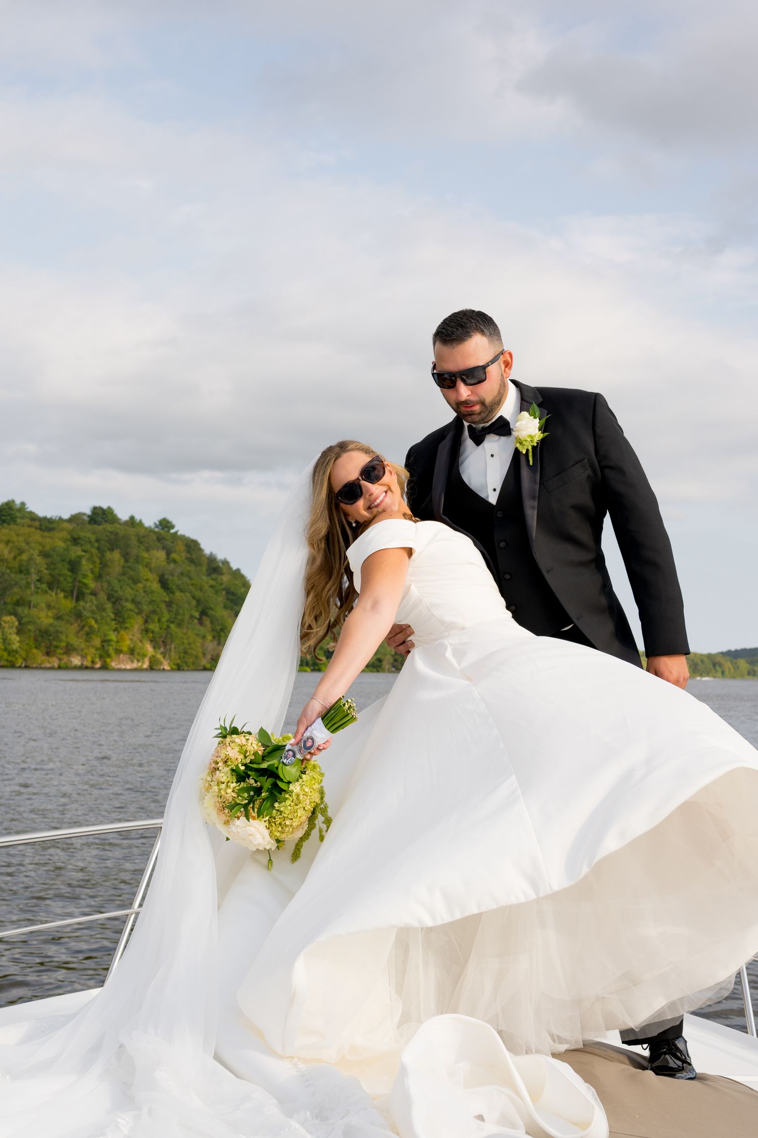 Wedding couple in formal attire posing on a boat deck against a scenic lake backdrop.