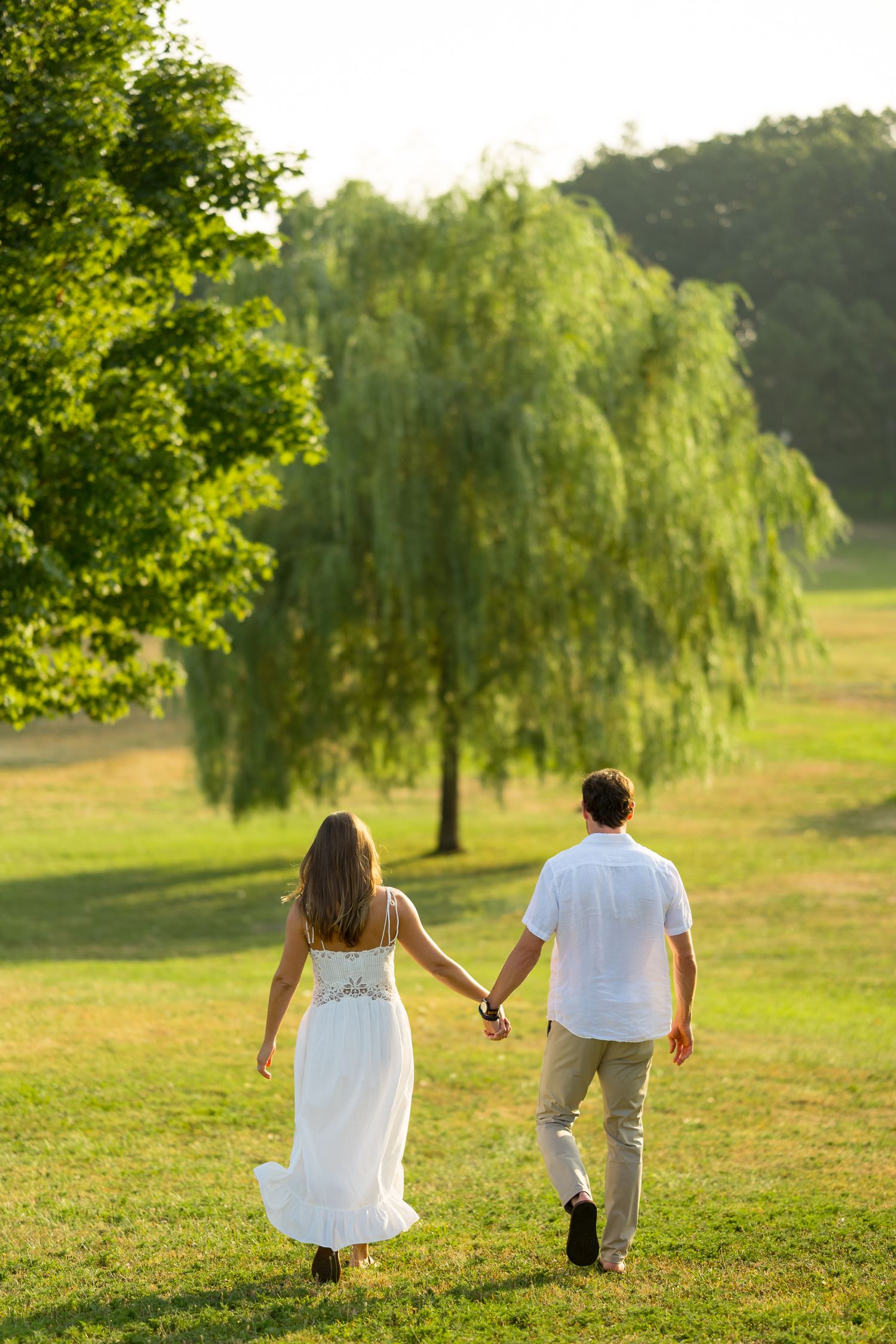 Two people in white clothing walk hand in hand across a sunlit grassy hill with willow trees in the background.