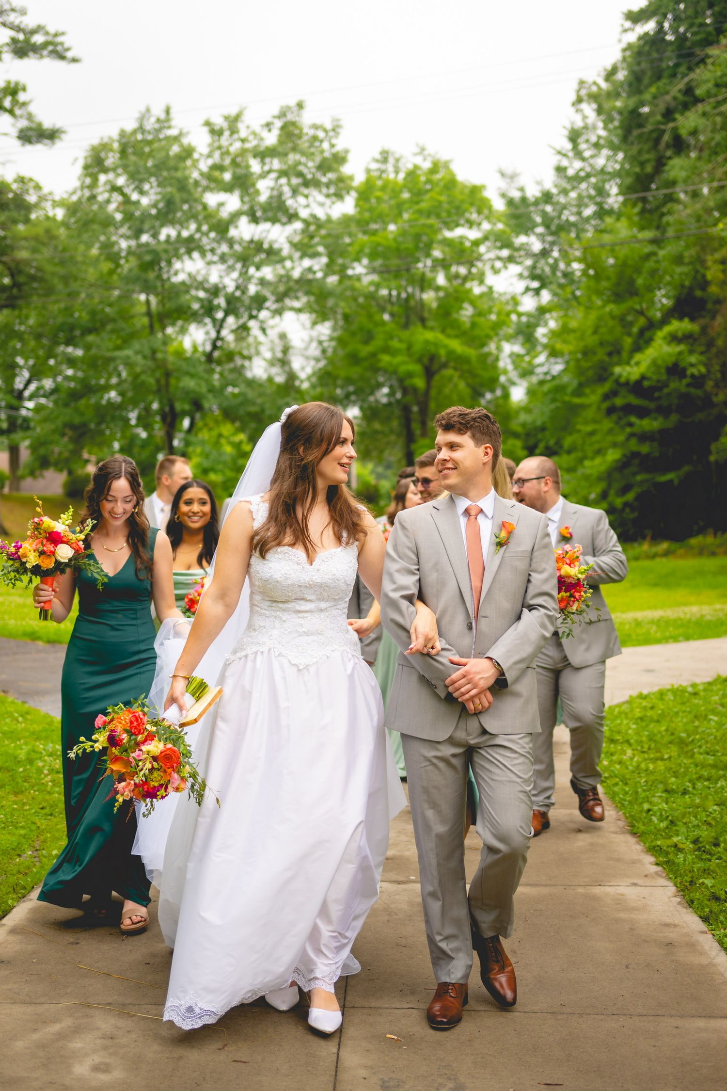Wedding party walks together on a path with bridesmaids in emerald dresses and groomsmen in gray suits.