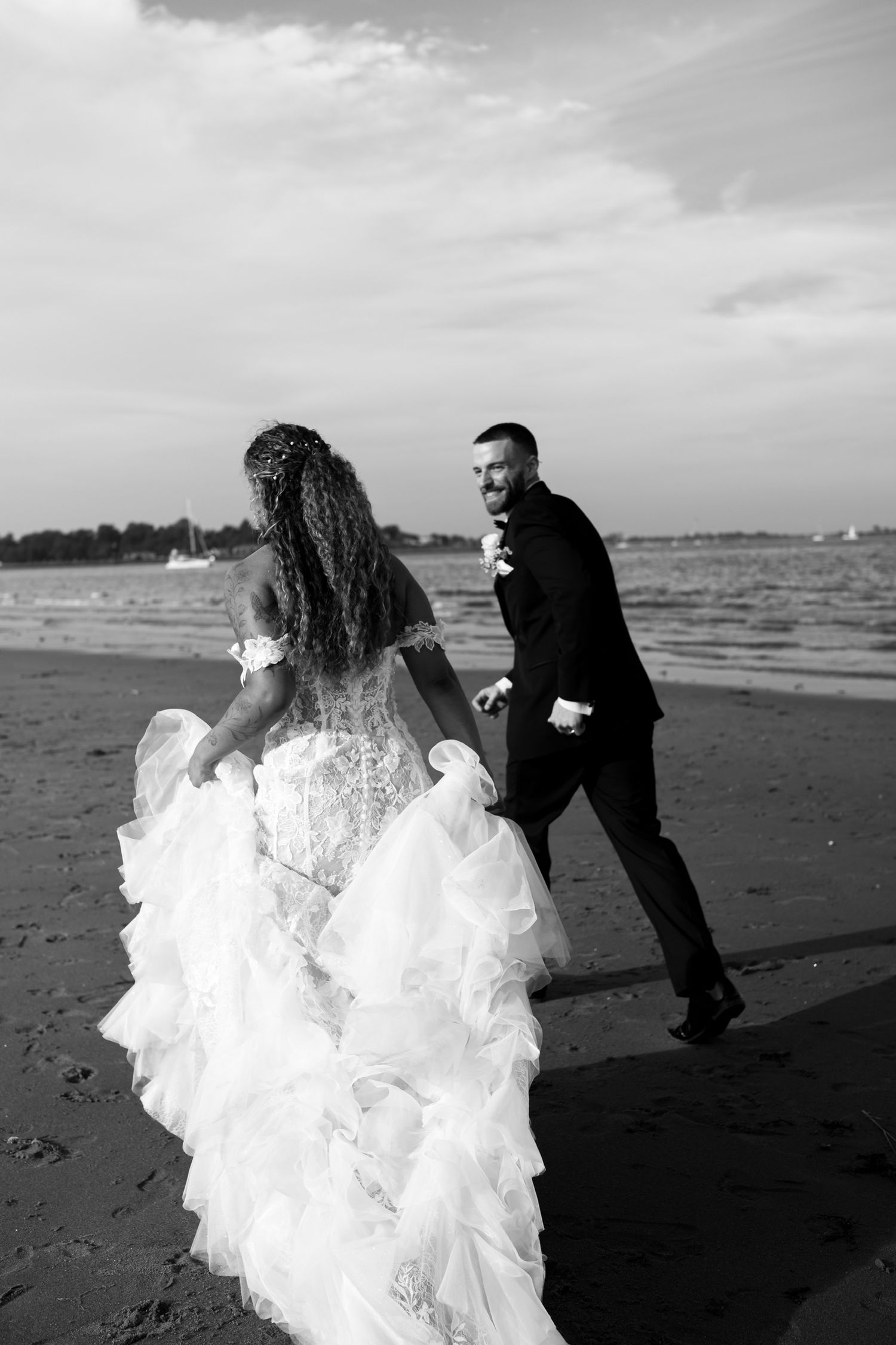 A couple in wedding attire runs playfully along a beach shoreline in black and white.