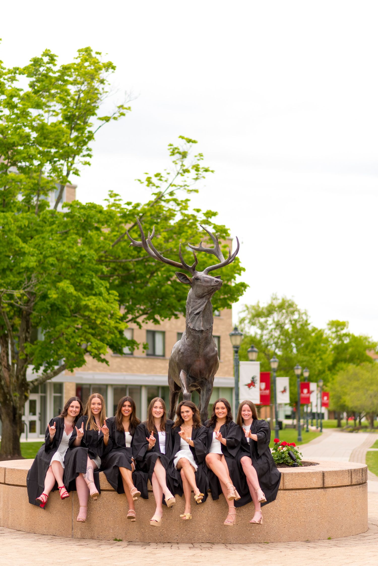 Fairfield University graduates pose together on a stone bench with stag statue on campus for graduation photos.