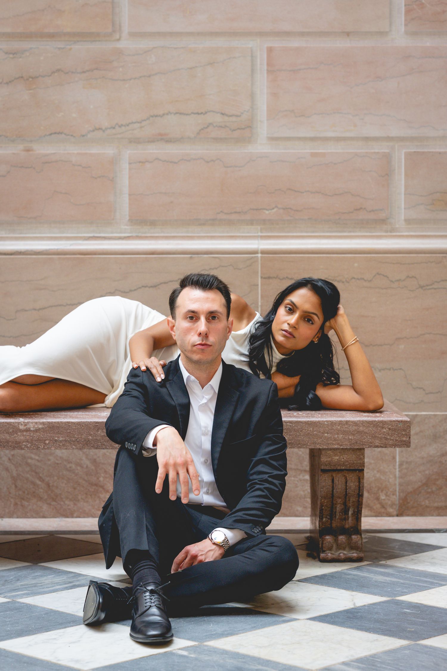 A couple relaxes casually on a checkered floor against stone walls in formal attire at Hartford City Hall.