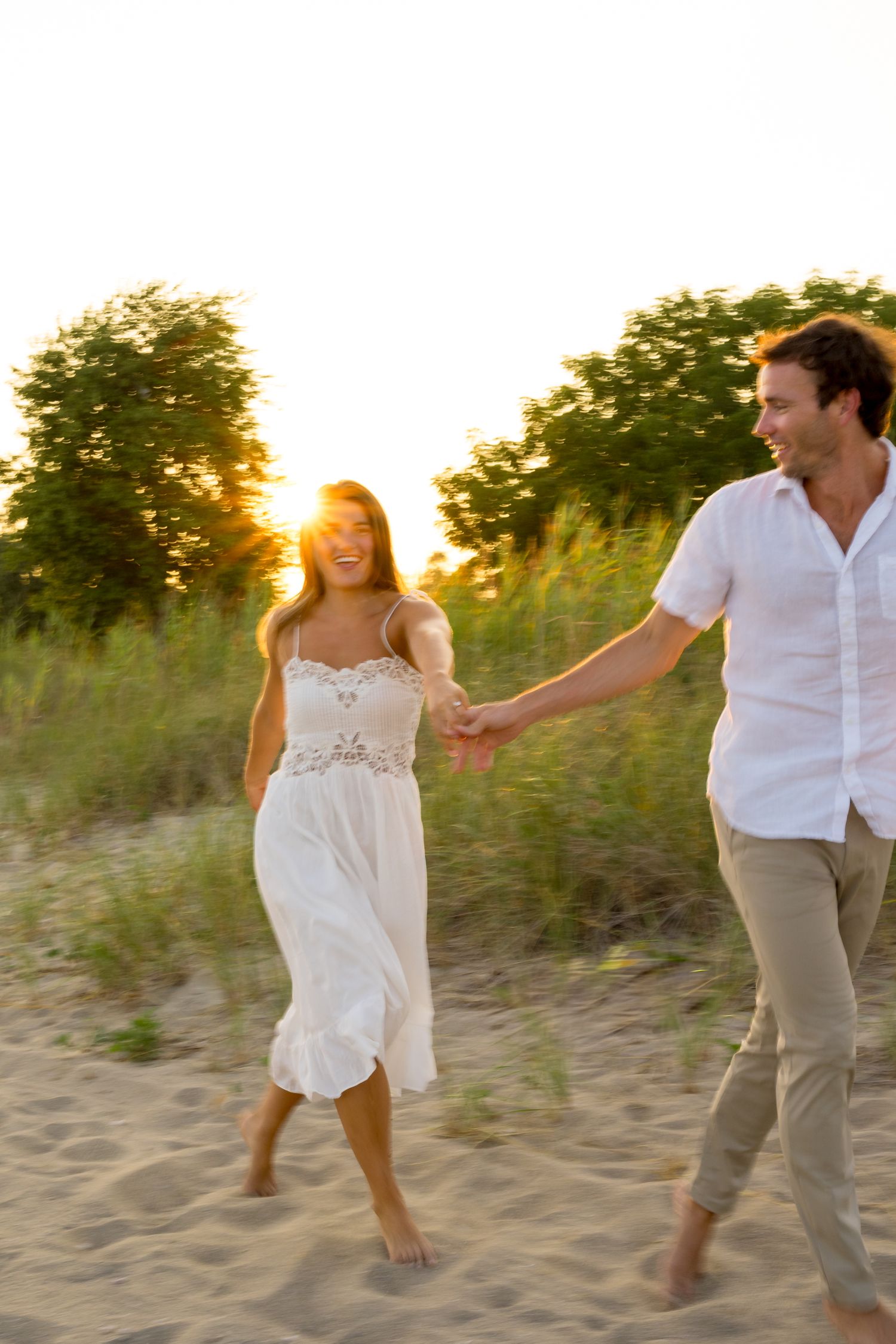 A couple plays and jumps together on a sandy beach at golden hour.