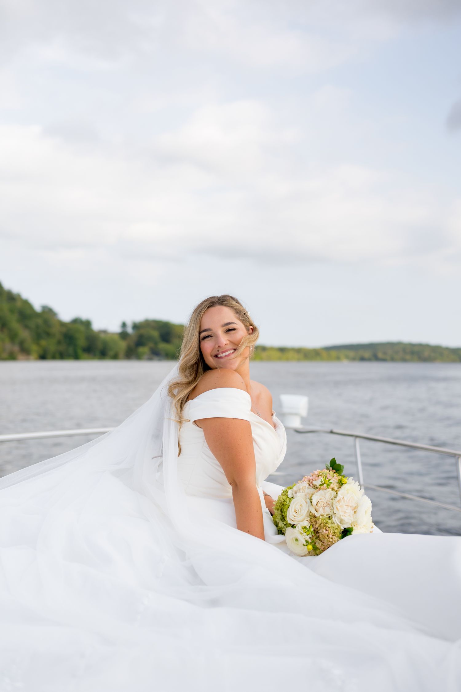 A bride in white dress poses joyfully on a boat deck with water and trees in background.