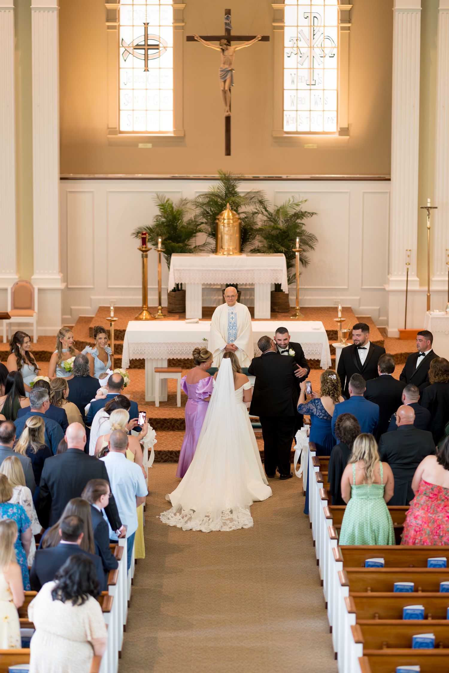 Wedding ceremony taking place in a traditional Catholic church with guests seated in wooden pews and floral decorations.