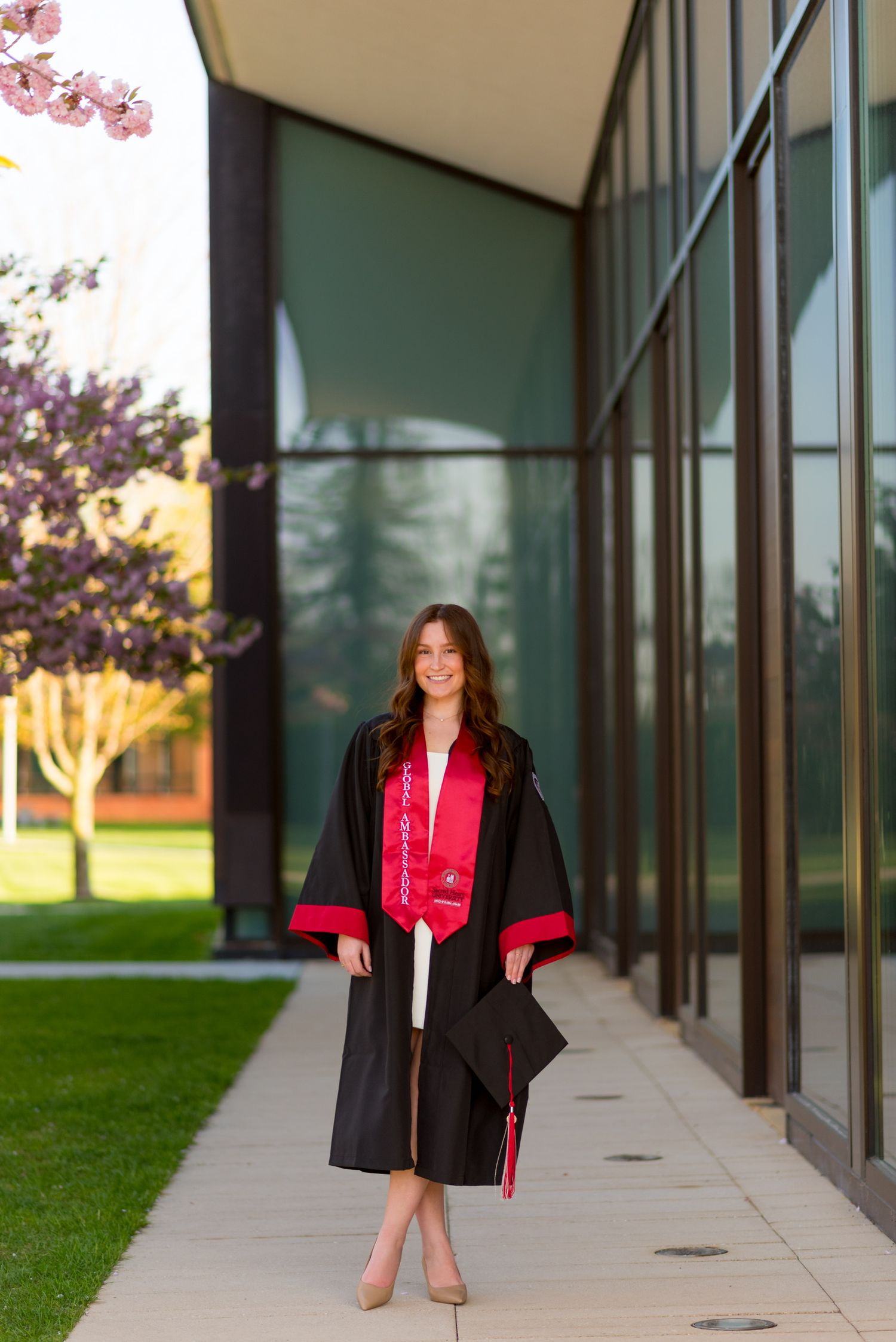 Sacred Heart University graduate in academic regalia posing outside modern building with glass windows.