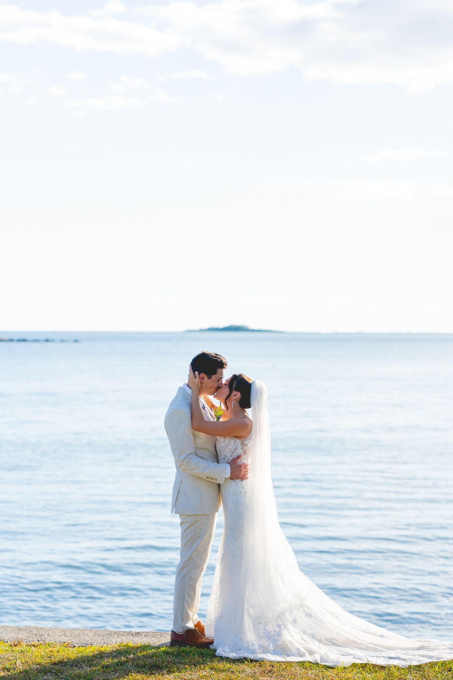 Wedding couple embracing by the ocean with bright blue water and sky in the background.