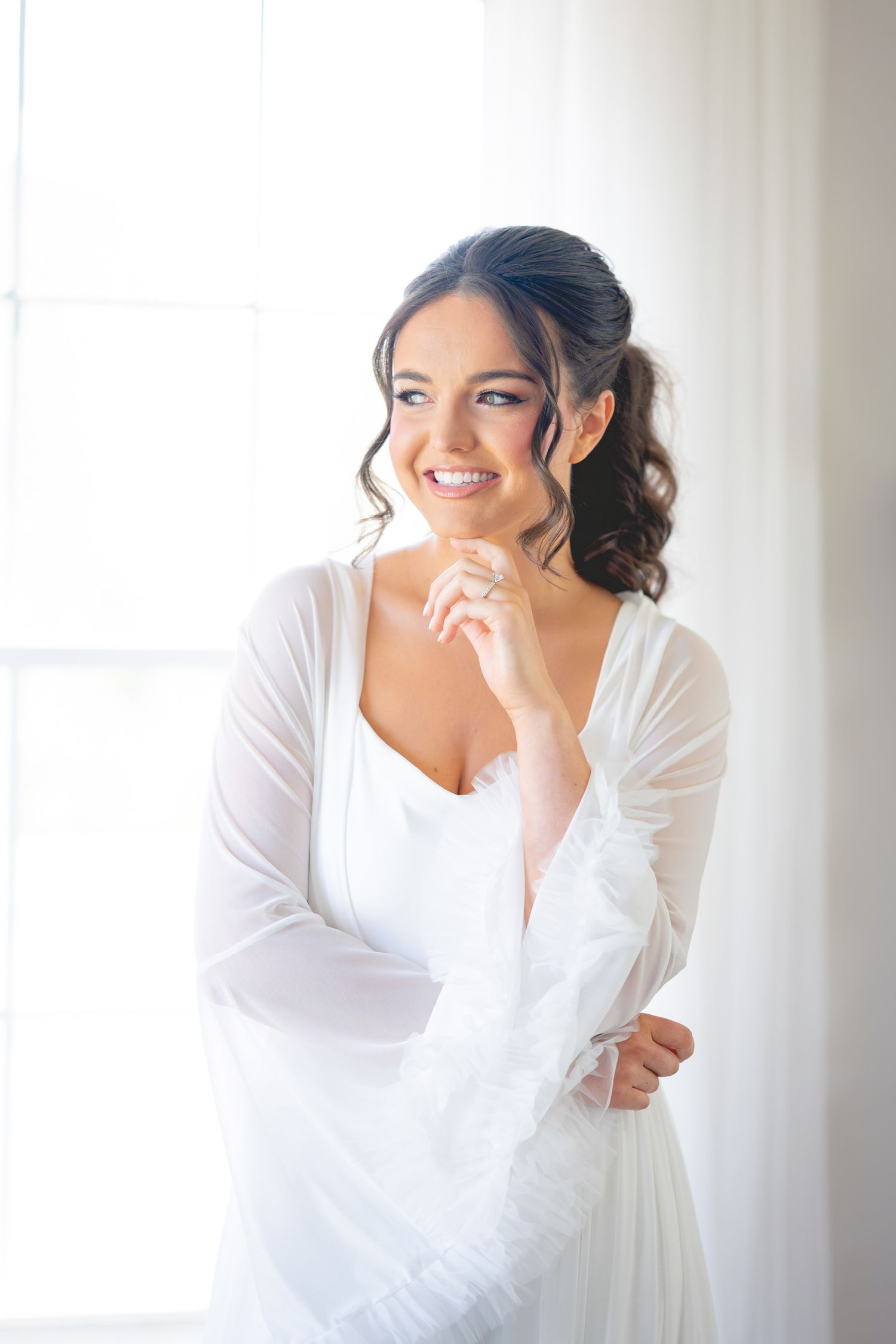 A bride in white laughs joyfully against a bright window background.