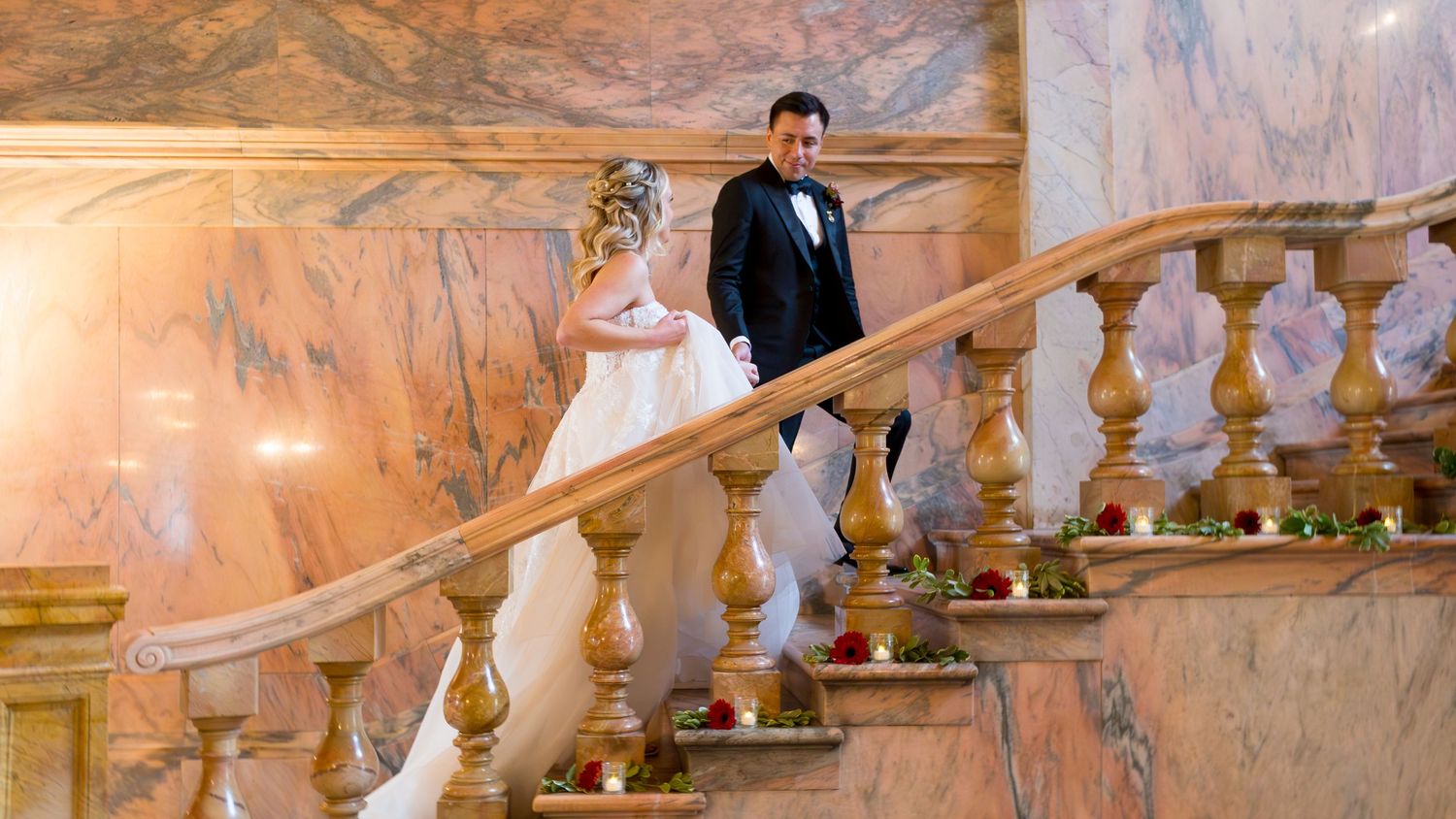 Elegant wedding couple descends grand marble staircase in ornate ballroom with peach-colored walls and decorative railings.
