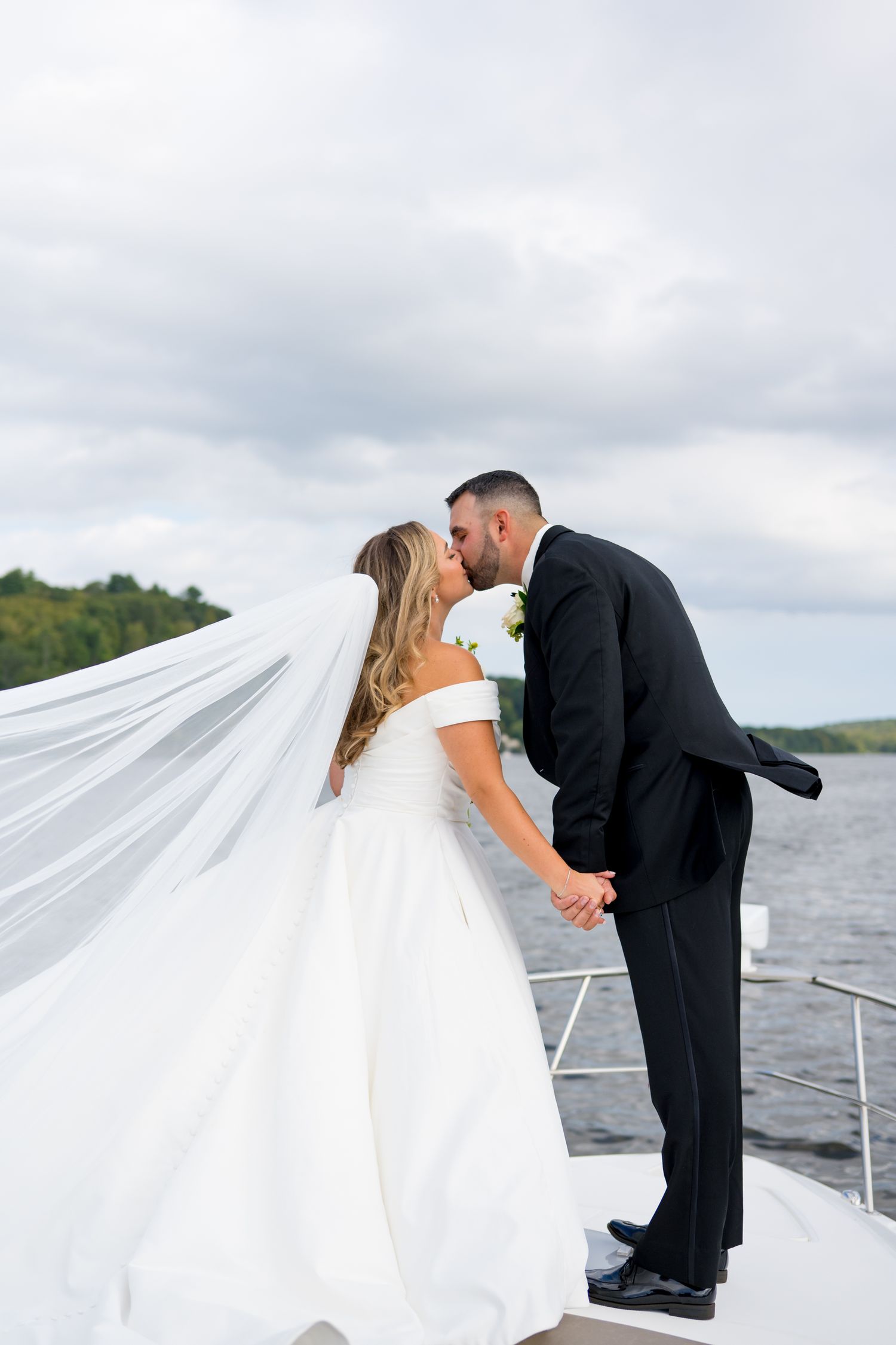 Romantic wedding couple sharing a tender kiss on a boat deck with flowing white veil against cloudy sky.