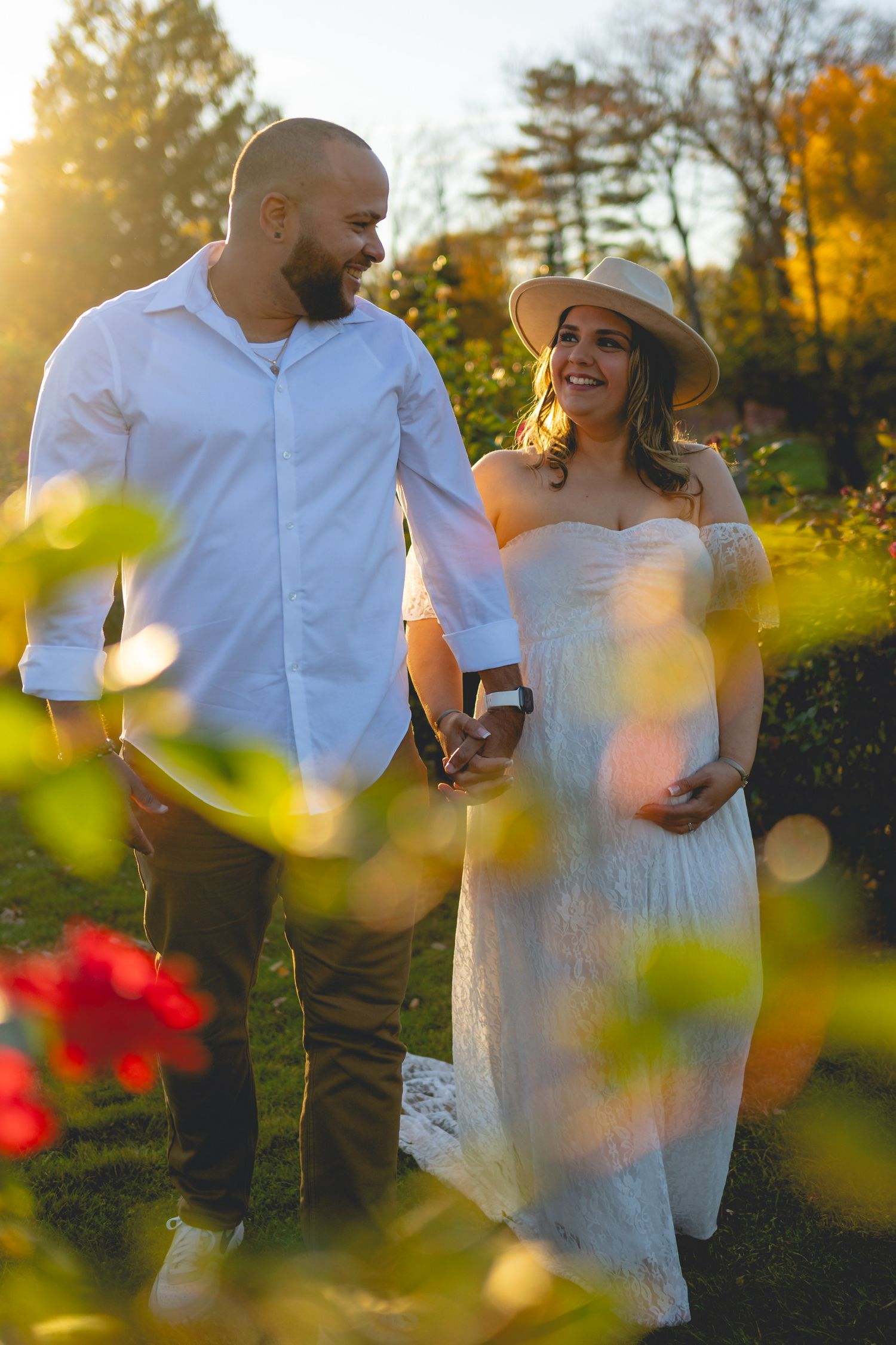 Romantic couple embraces in a sunlit garden with autumn foliage and red flowers in the foreground.