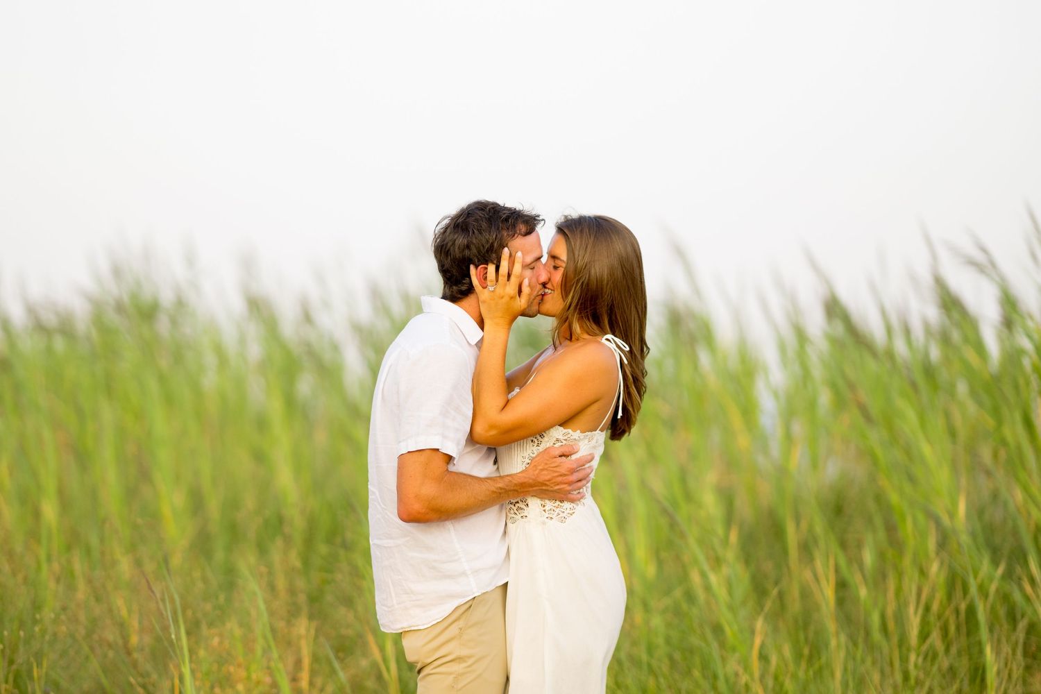 A romantic couple embraces on the beach with tall grass swaying in the foreground.