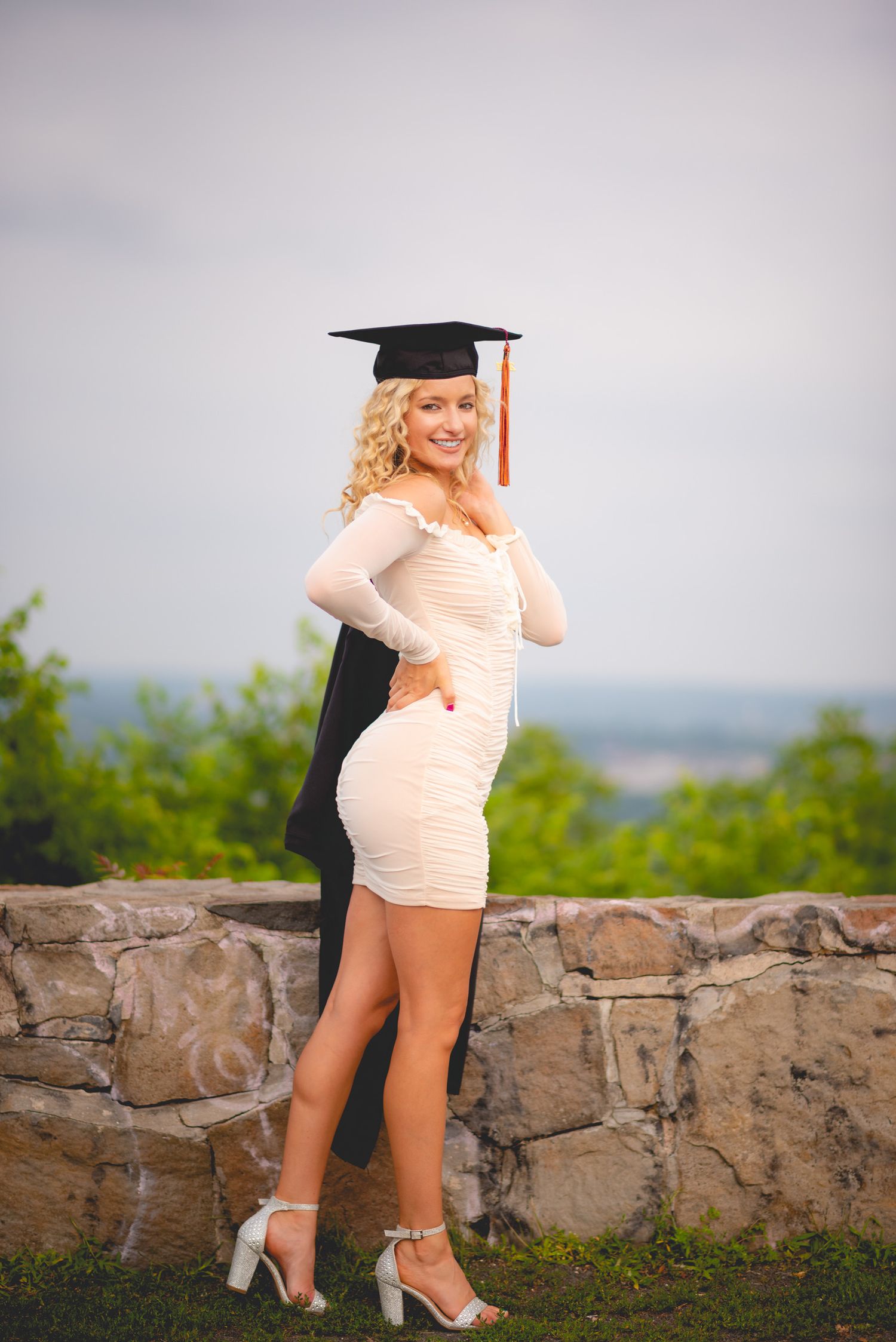 Graduate in white dress and cap poses on stone wall with scenic overlook for graduation photos.
