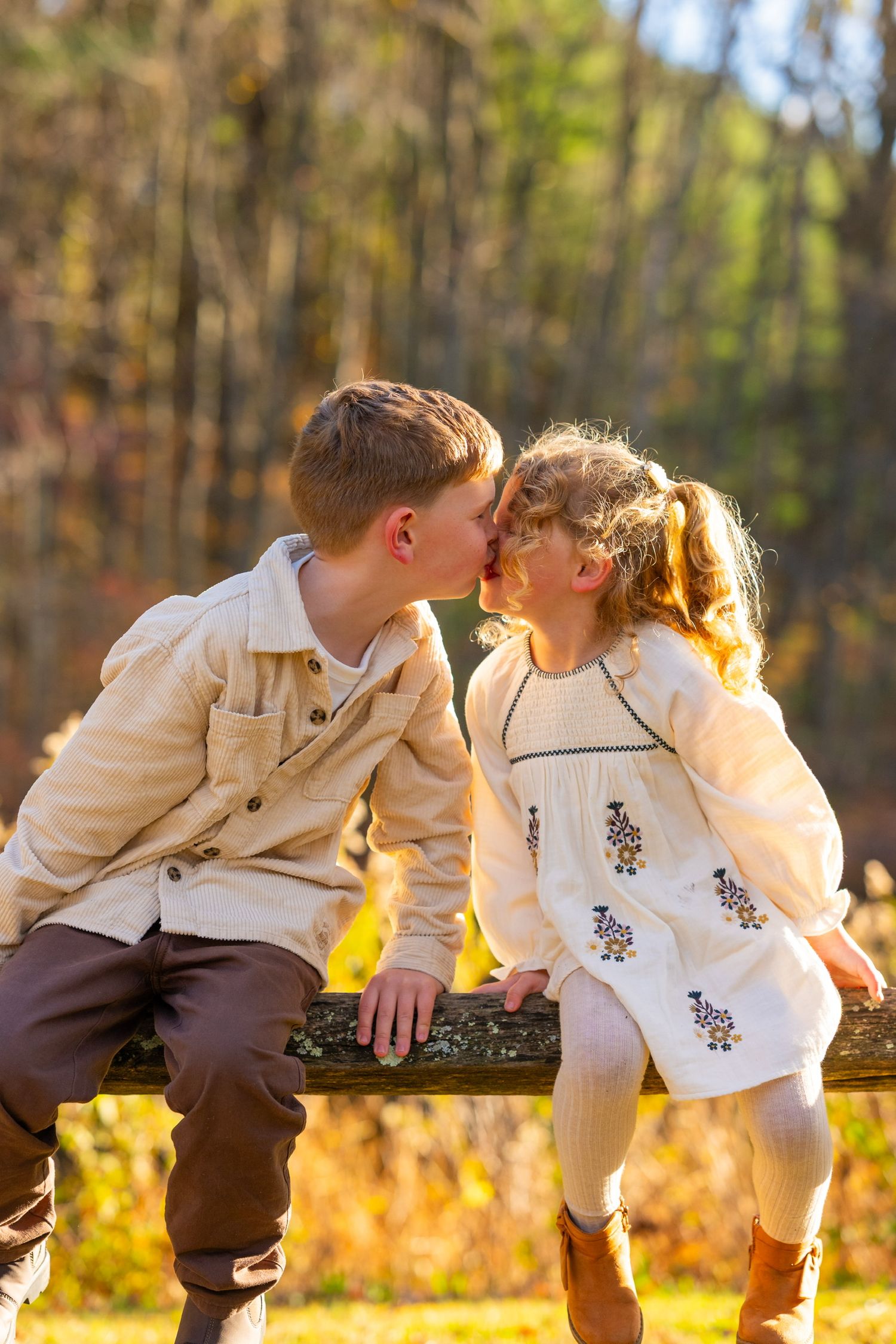 Two young children in matching white outfits share a sweet moment on a wooden fence for family photoshoot.