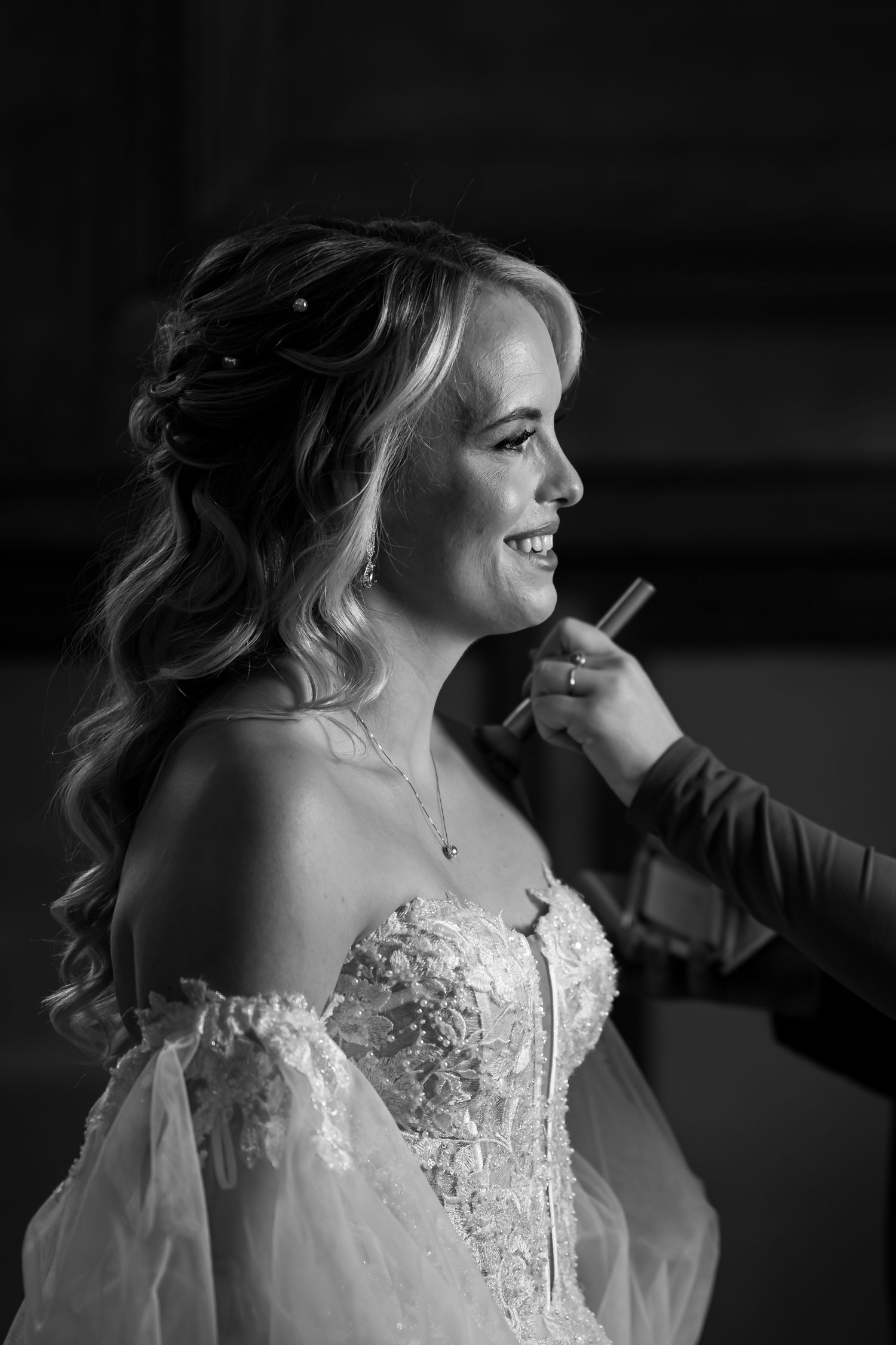 Black and white photo of a bride in a lace wedding dress with sheer sleeves smiling during wedding preparations.