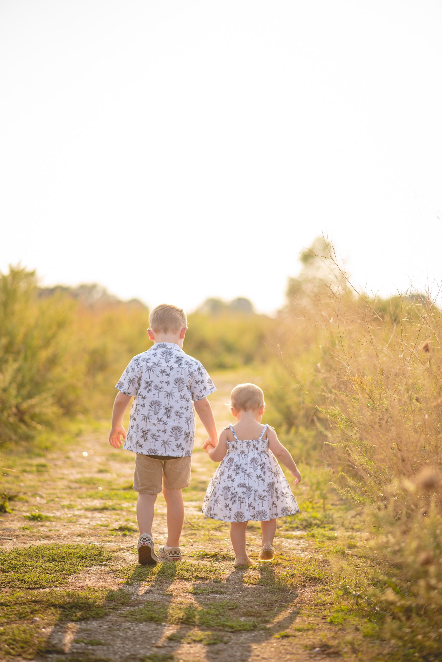 Two small children walking together on a sunlit path through tall grass.