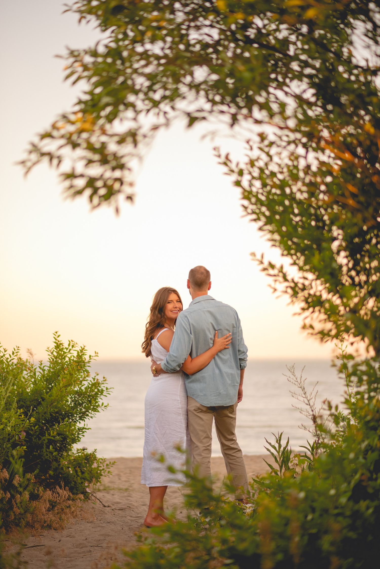 A romantic couple embraces on a beach at sunset with trees framing the scene.