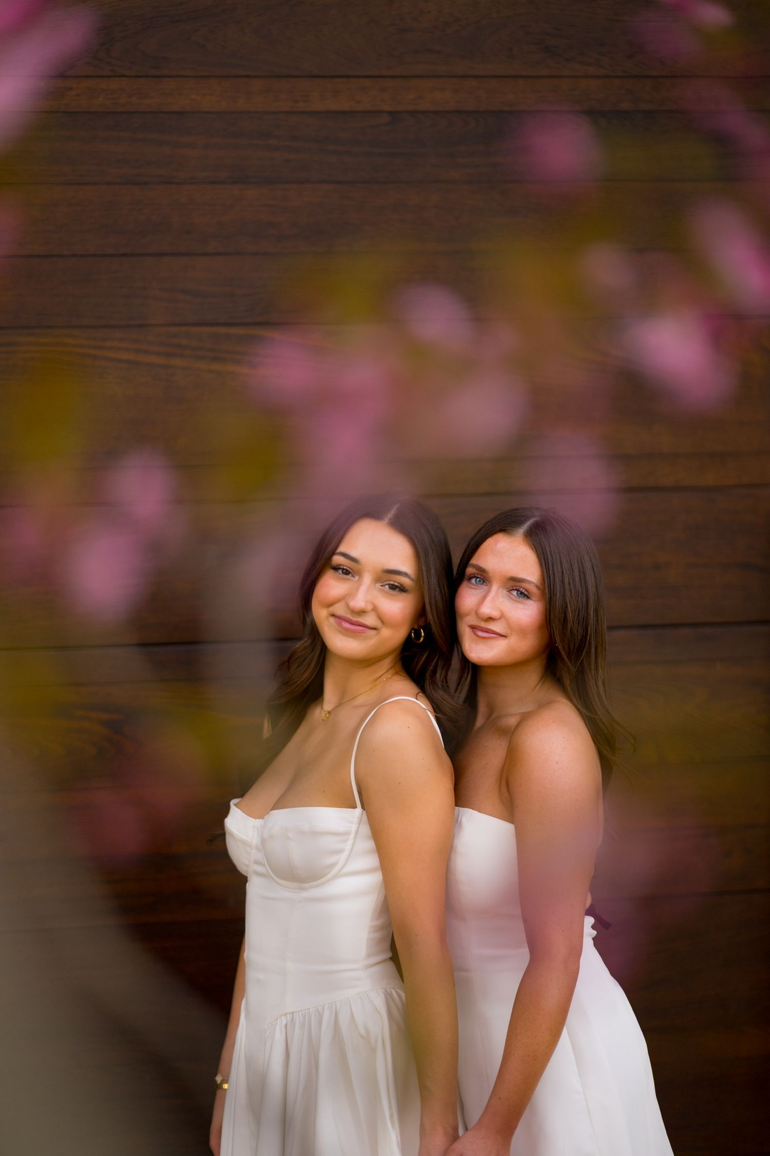Two Sacred Heart University graduates in white dresses pose together against wooden wall with pink blossoms in foreground.