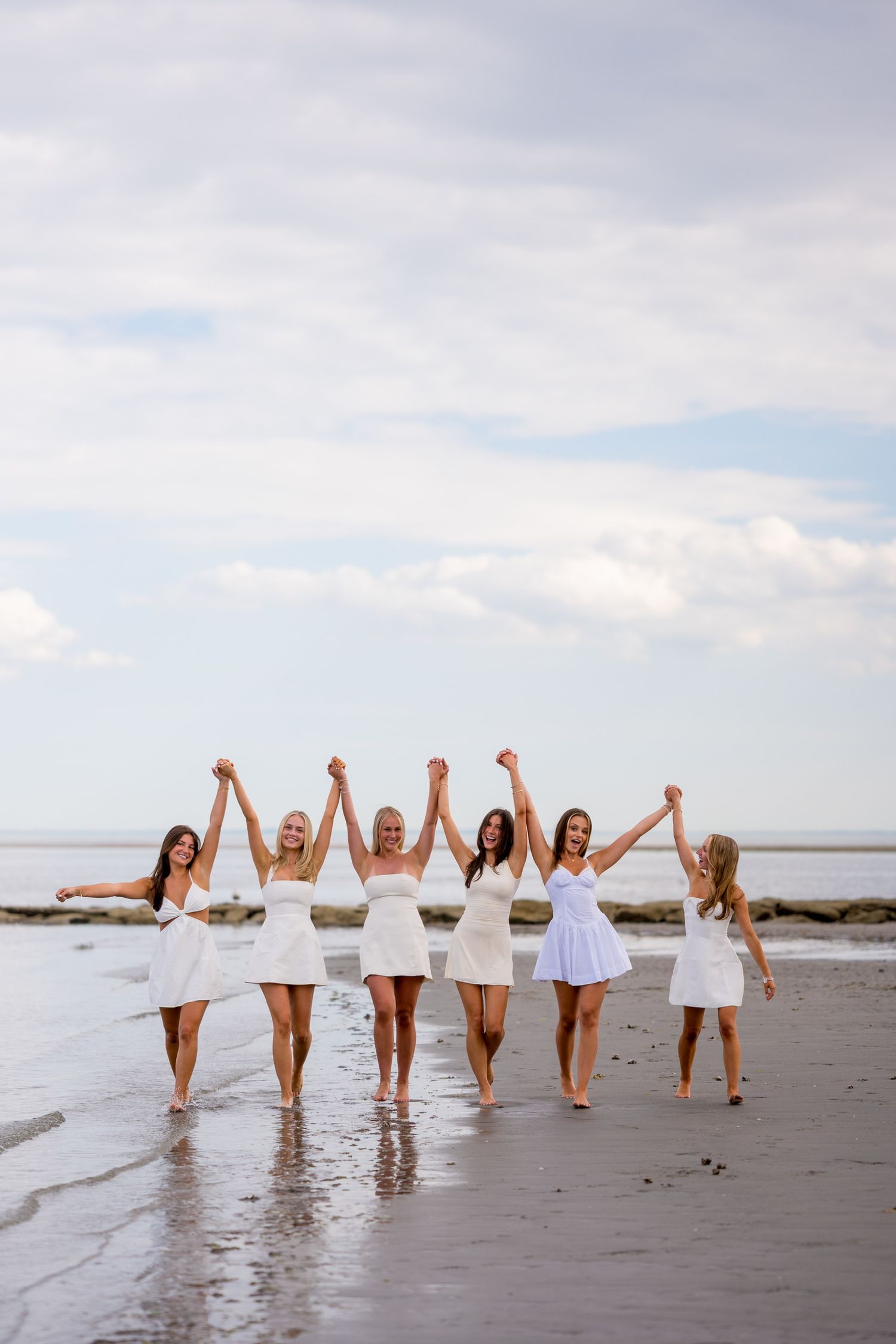 Fairfield University graduates in white dresses walking along a beach shoreline for graduation photos.
