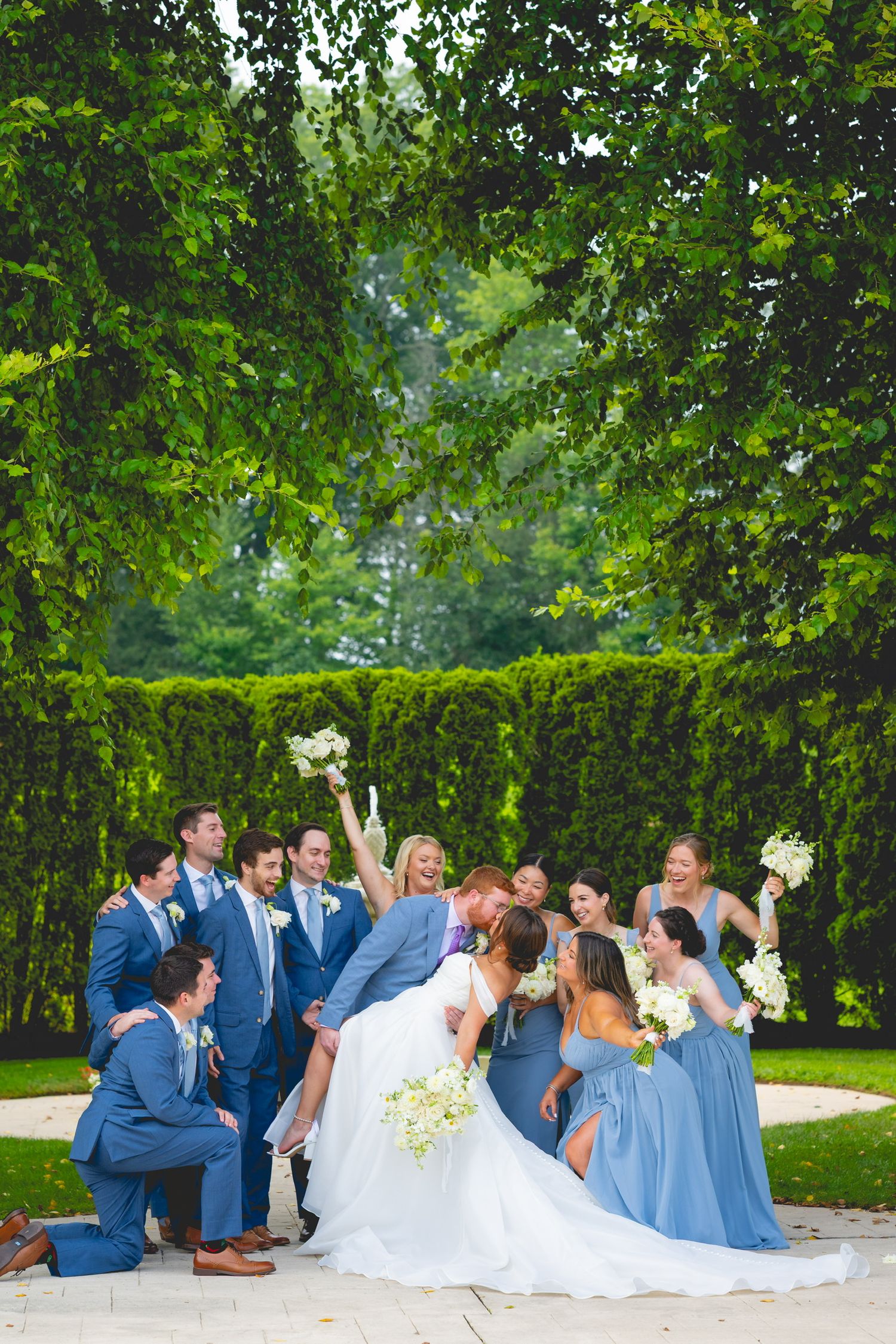 Wedding party in navy blue and light blue attire lounges on grass under green trees at the Waterview in Connecticut.