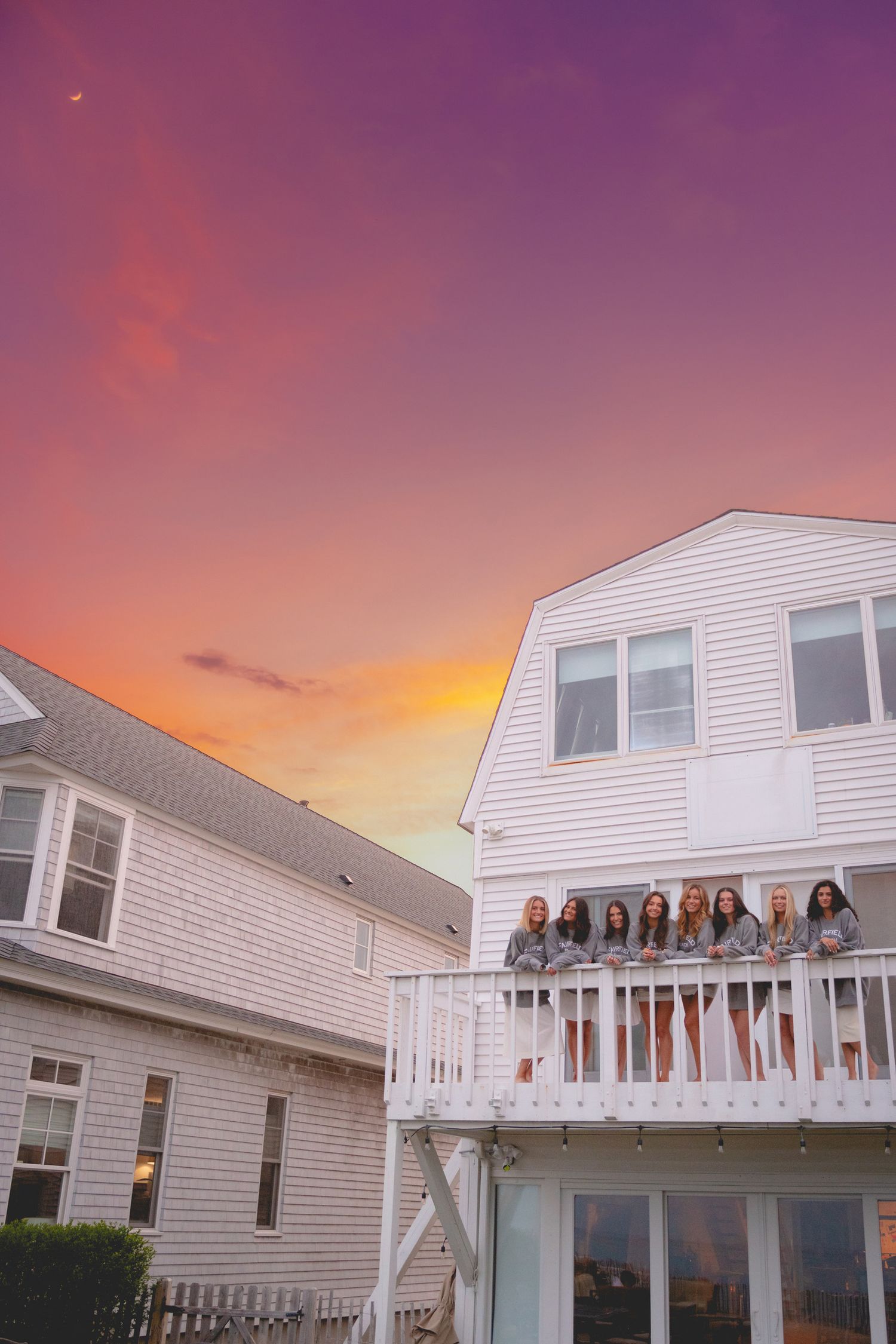 Fairfield University graduates in white dresses pose together on a beachfront deck during a vibrant sunset.