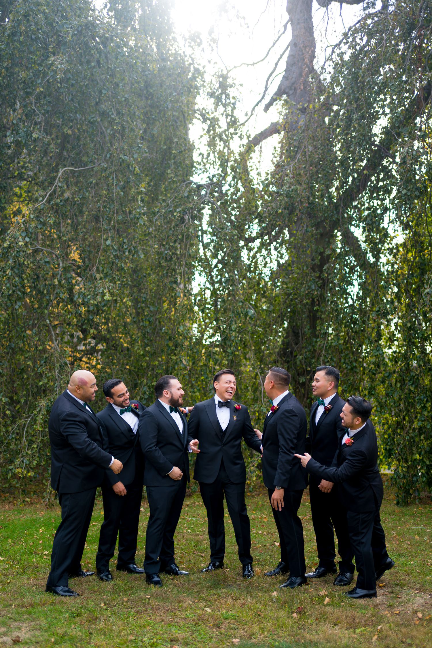 A group of men in black tuxedos and bow ties share a candid moment under weeping willow trees.