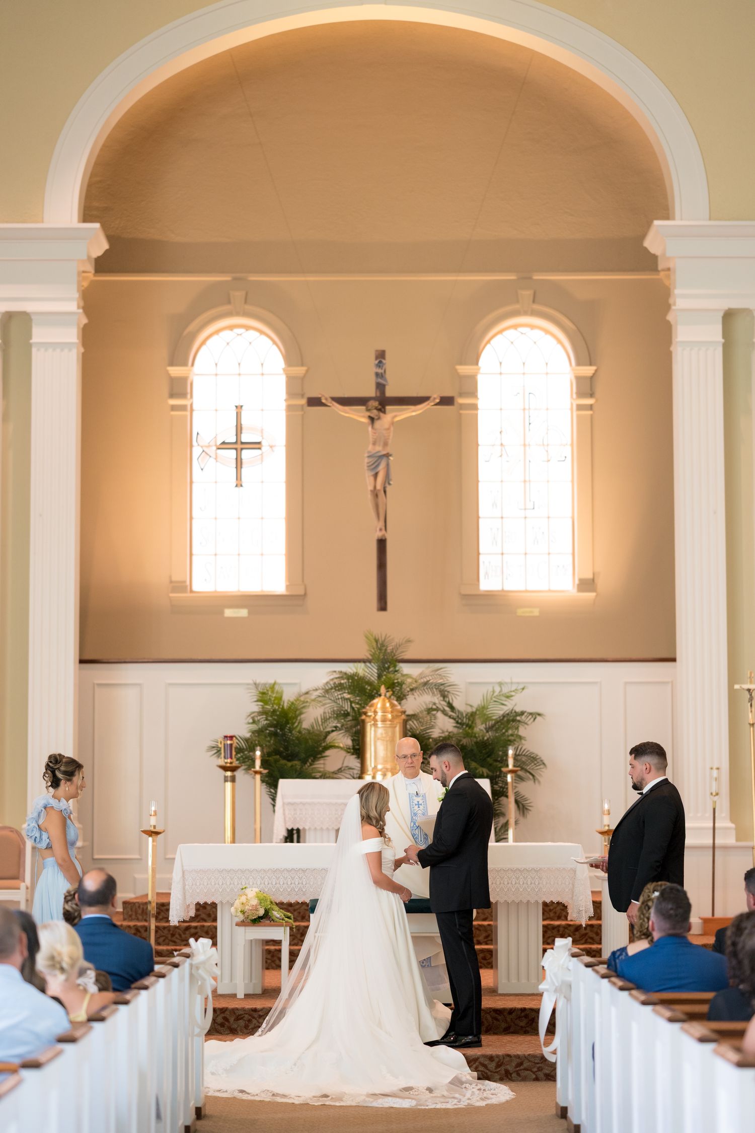 Catholic church wedding ceremony in progress with white pews and arched windows in a traditional sanctuary setting.