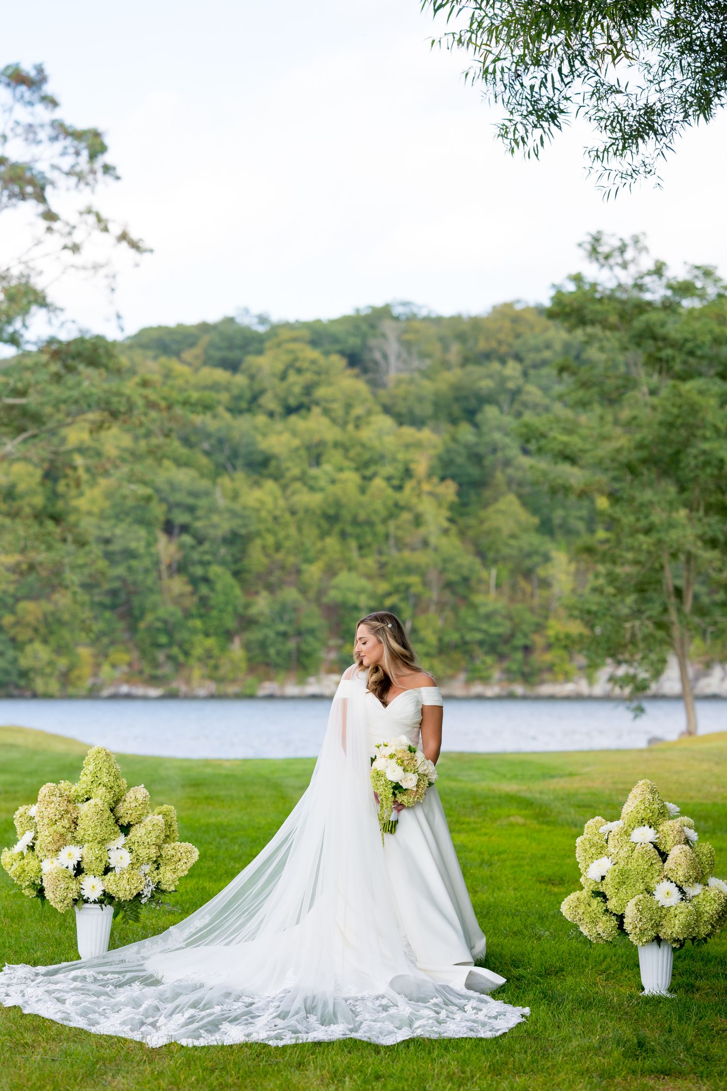 A beautiful white wedding dress spreads across green grass near a lake with hydrangea flowers and trees in the background.