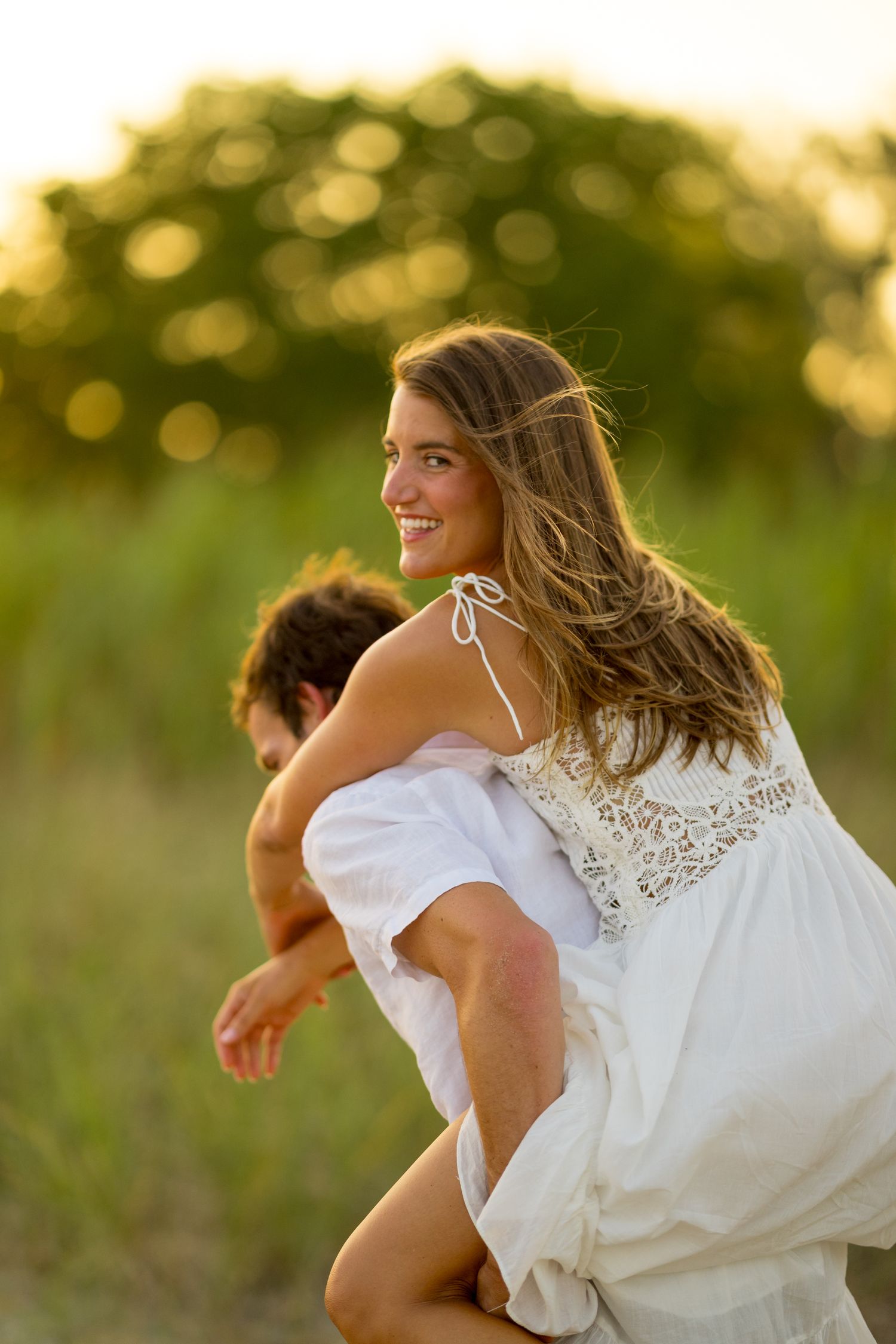 A couple laughs while spinning together on a beach at sunset with golden bokeh lighting.