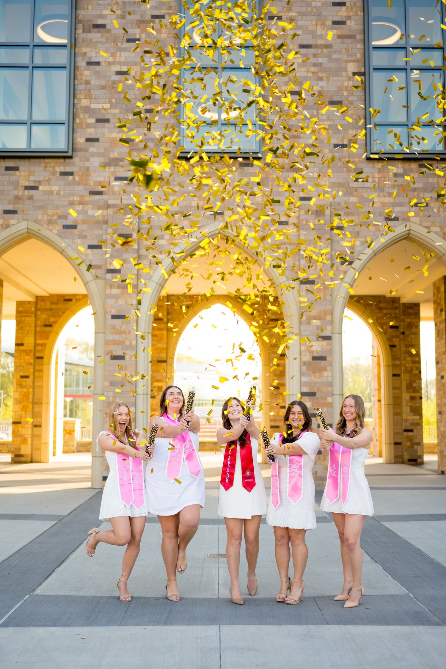 Sacred Heart University graduates throwing gold confetti in front of a brick building with arches during celebration.