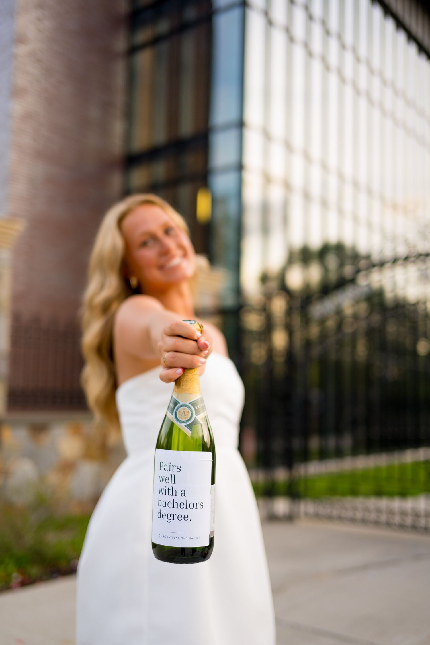 Sacred Heart University graduate in white dress holds champagne bottle while celebrating outdoors.