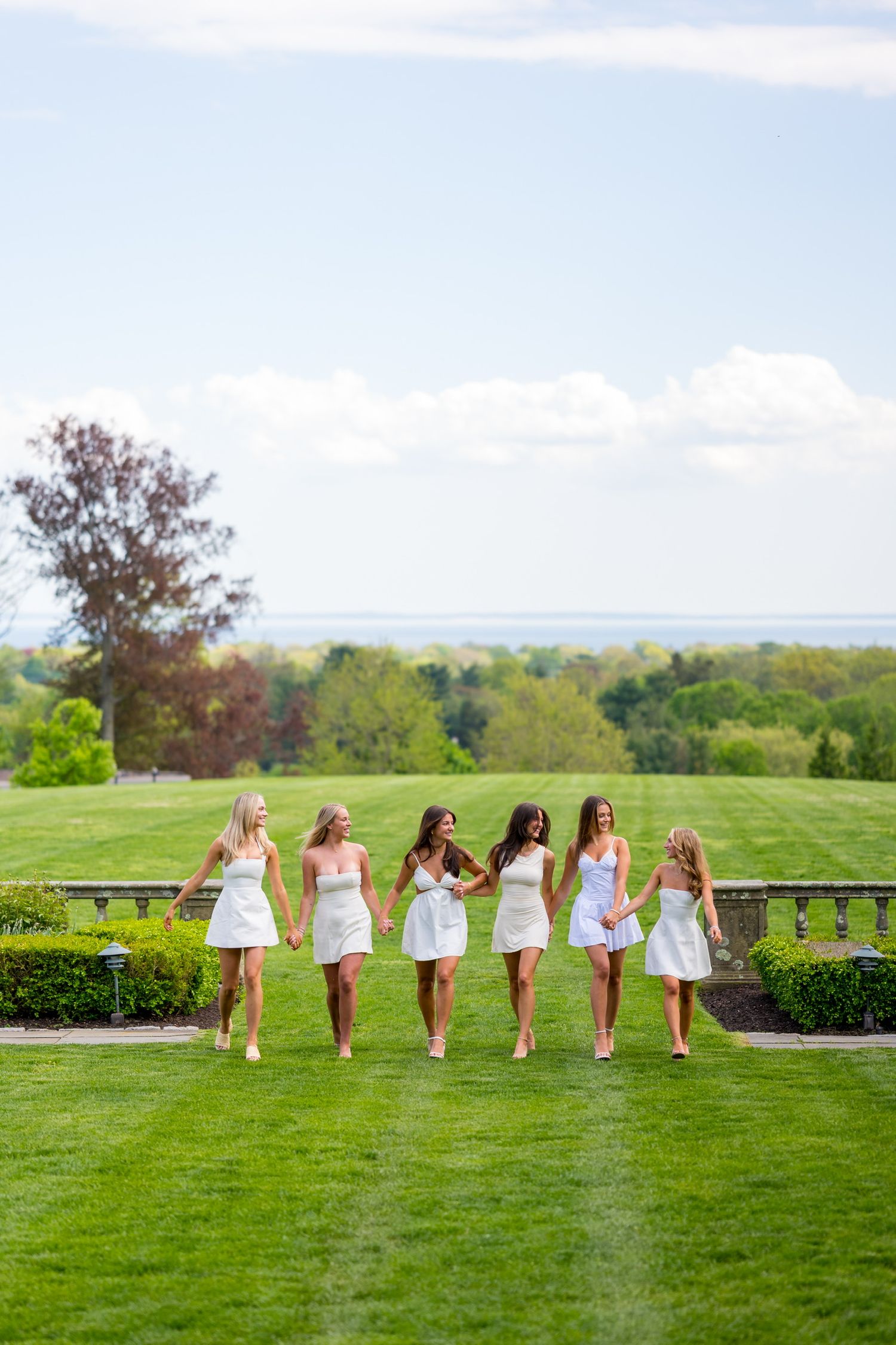Fairfield University graduates in white dresses walks across manicured lawn with scenic landscape view.