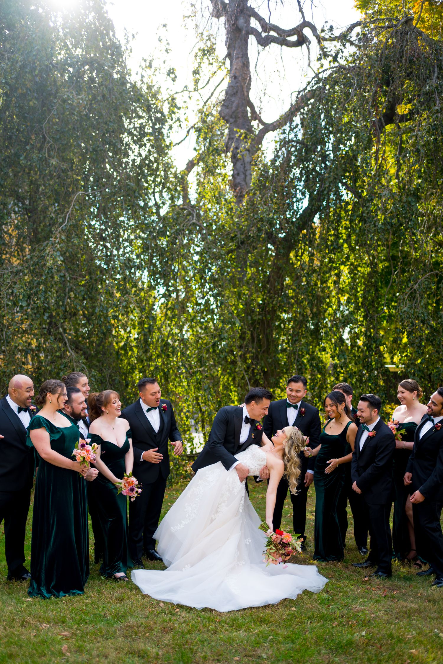 Wedding party in formal attire stands in a row under sunlit trees during an outdoor celebration.