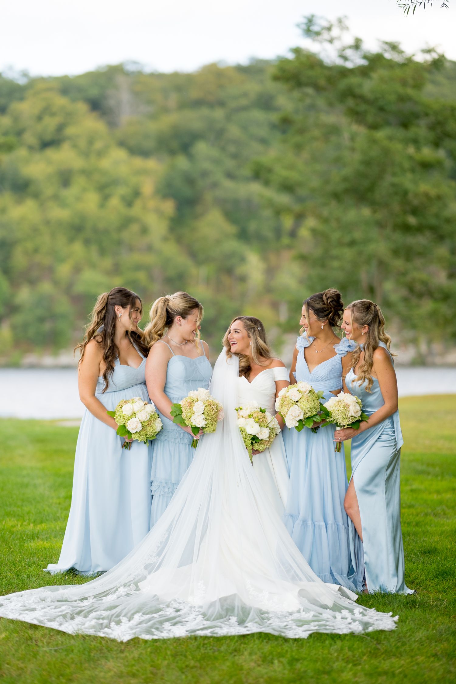 Wedding party in light blue bridesmaid dresses and white bridal gown pose together on grassy lawn by lake.
