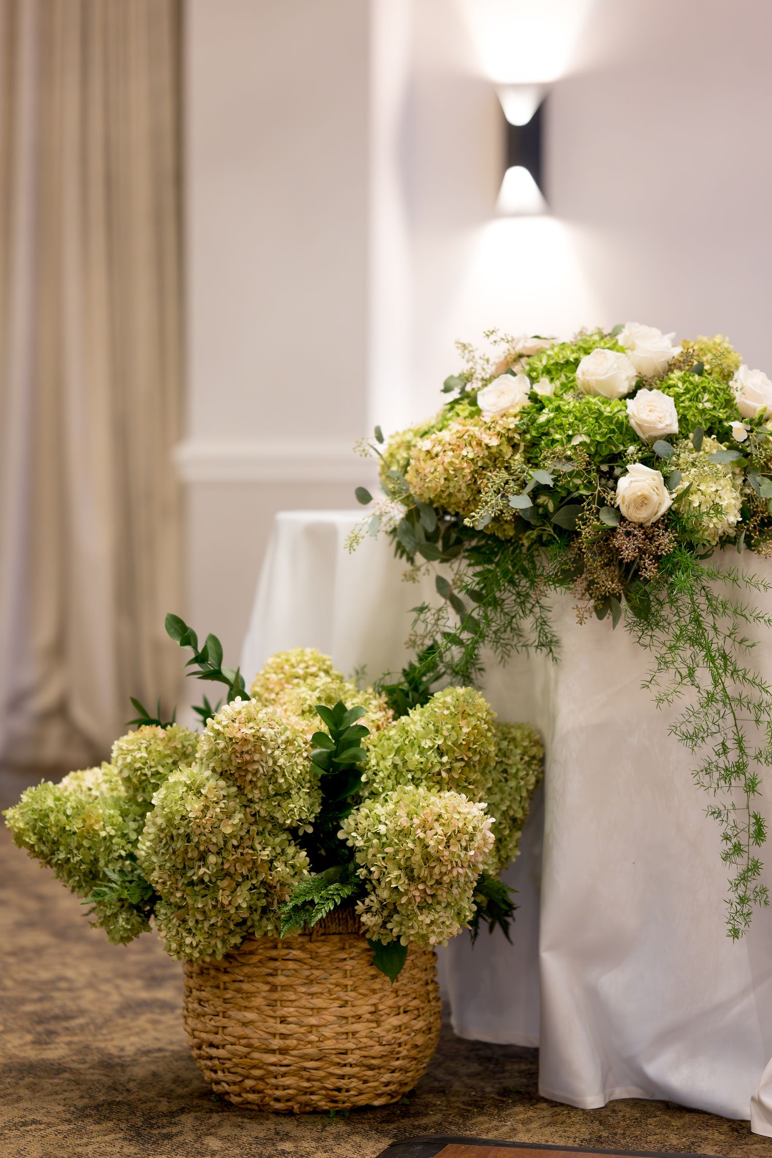 Elegant floral arrangement with white roses and green hydrangeas in a woven basket against a light curtain backdrop.