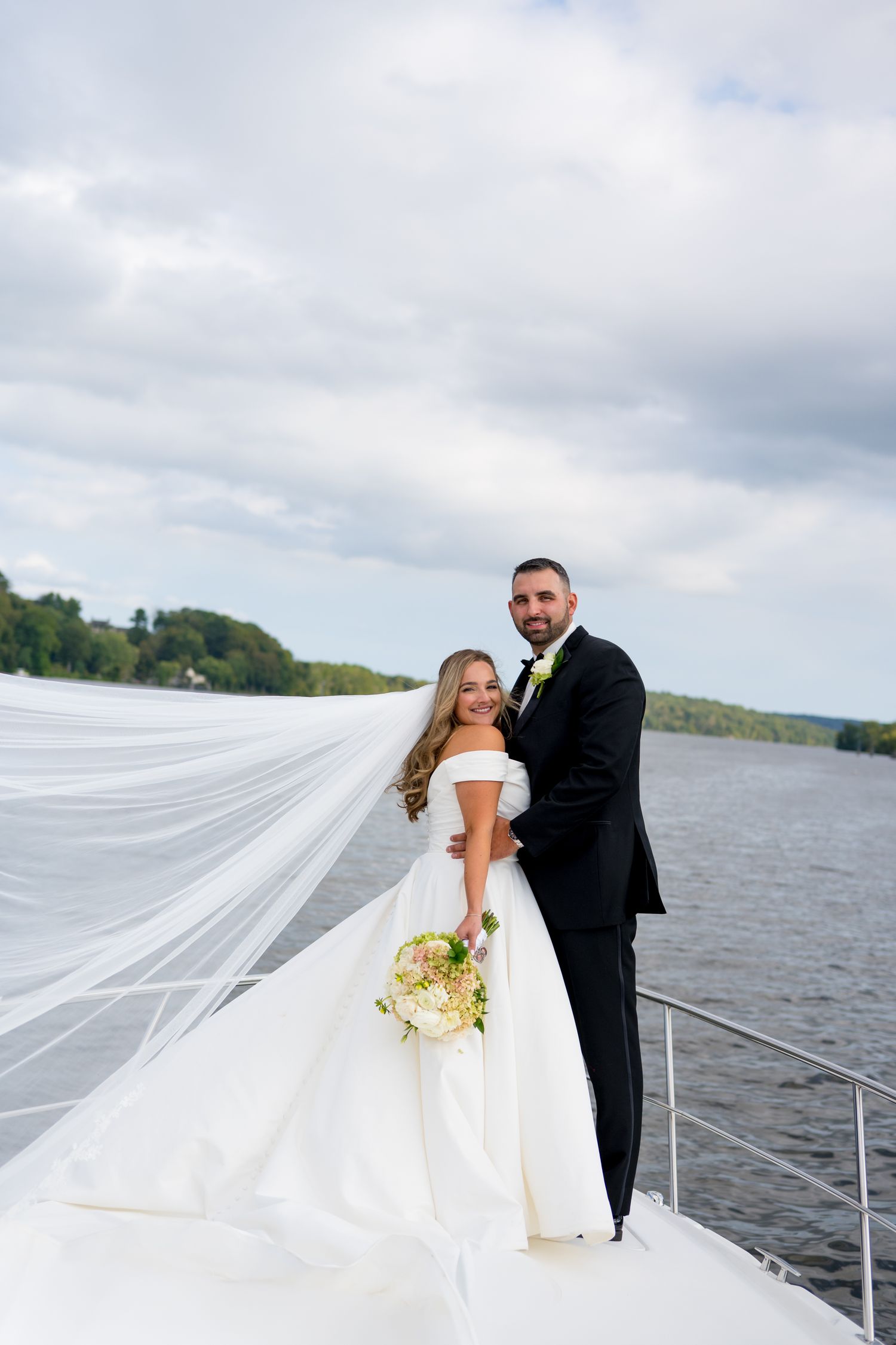 A couple poses on a boat deck with a flowing veil against a waterfront backdrop.