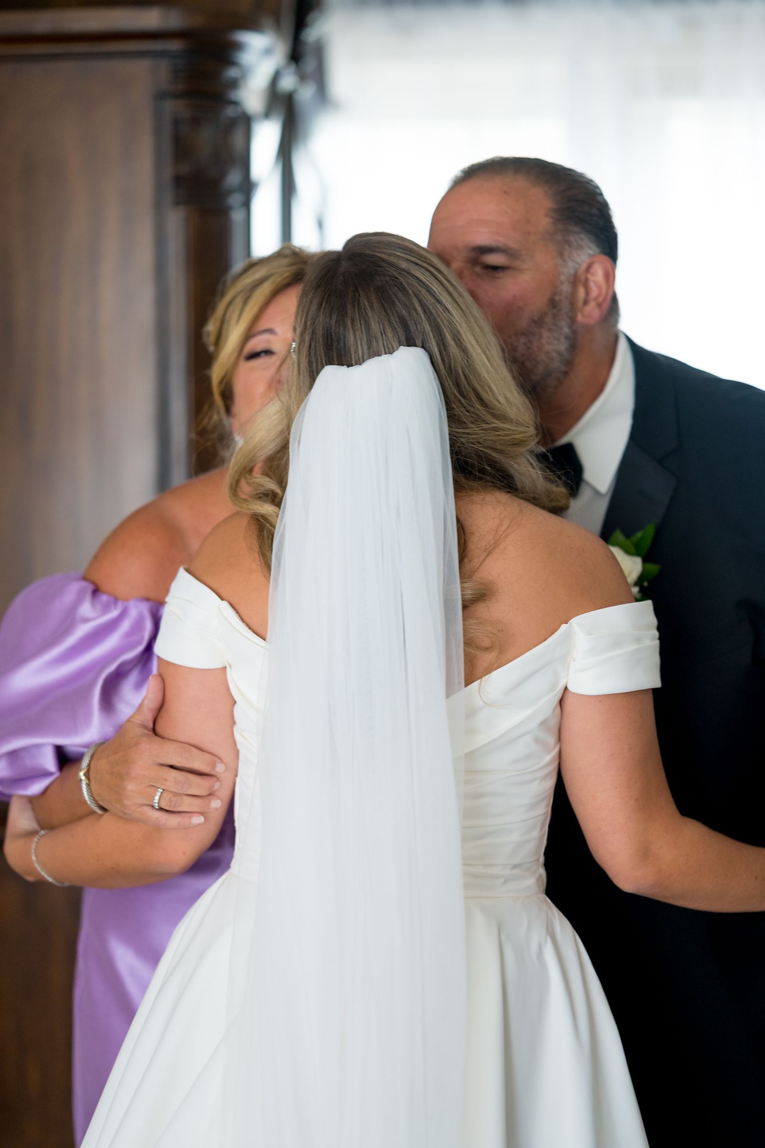 A bride in an elegant off-shoulder white wedding dress receives kisses from two guests at her wedding celebration.