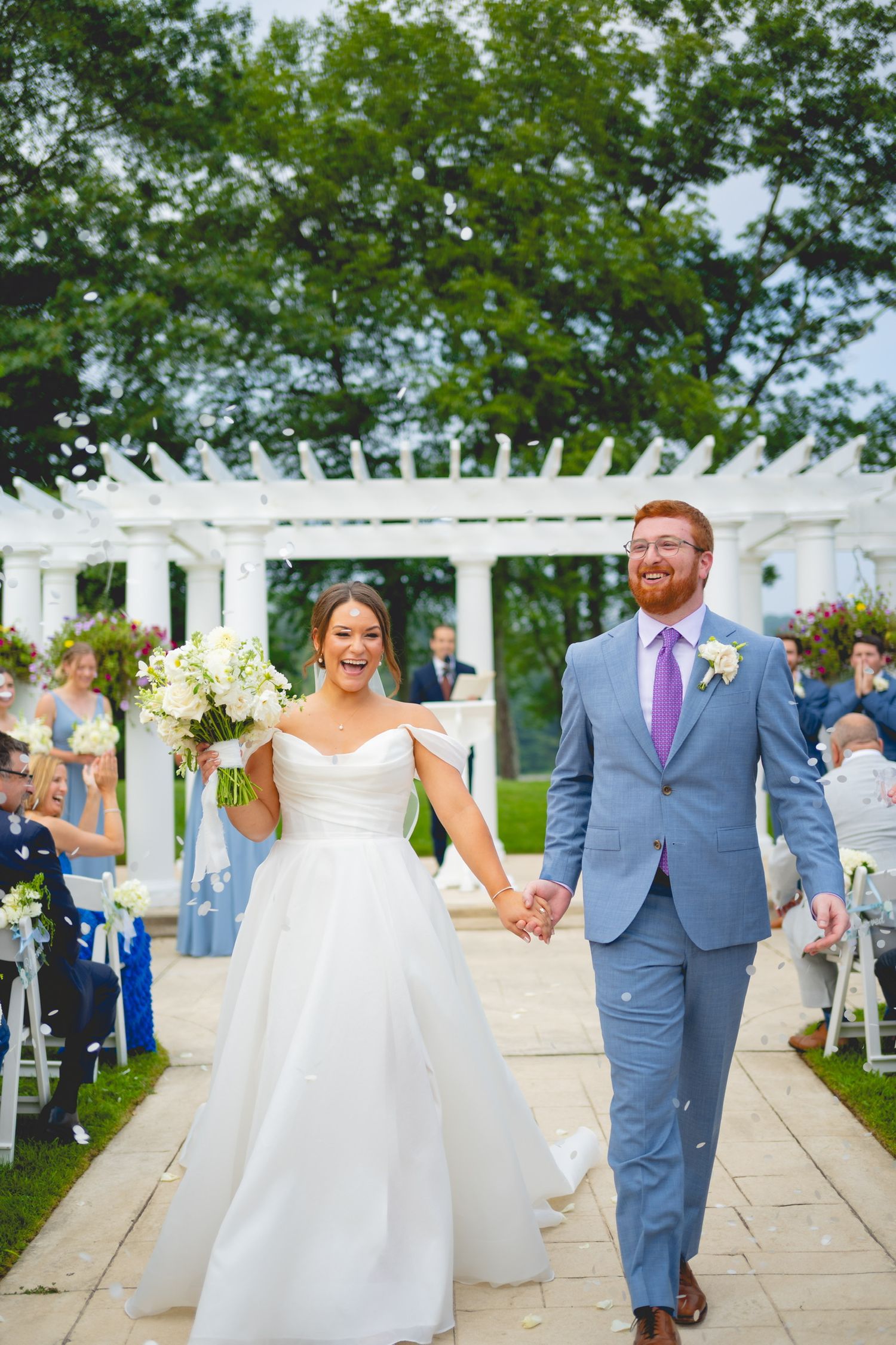 Newlyweds walking down aisle under white pergola surrounded by garden flowers.