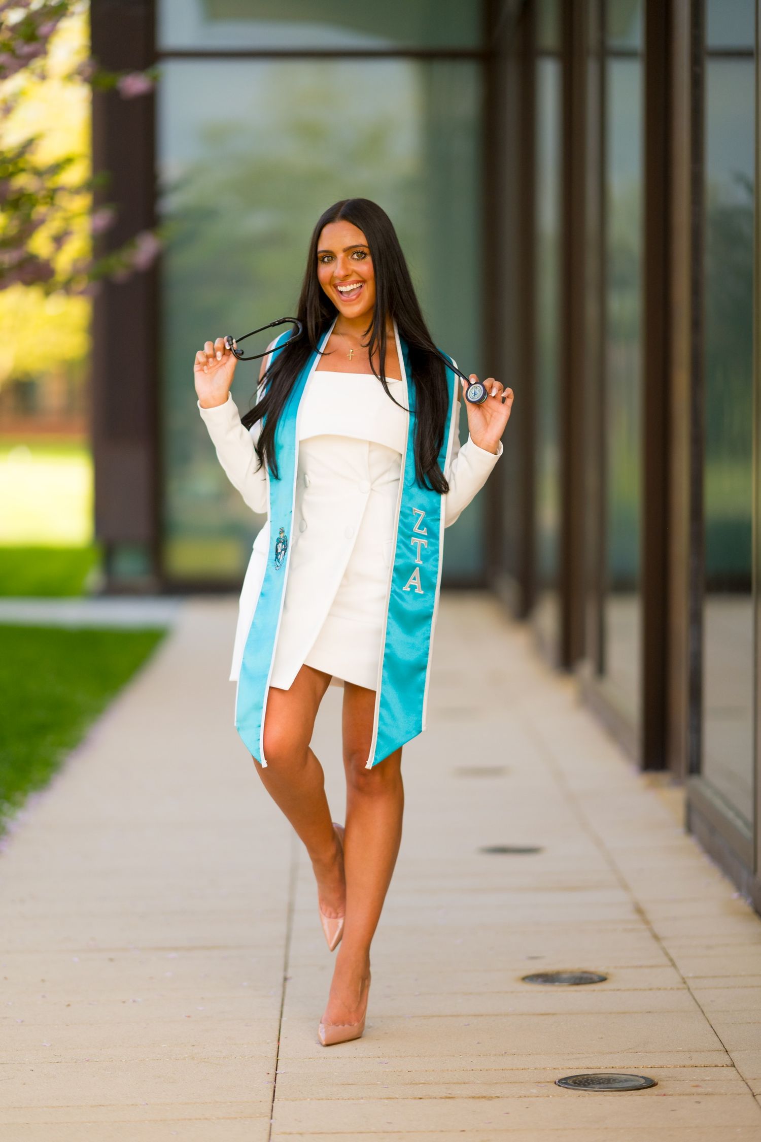 Sacred Heart University graduate in white dress and turquoise graduation sash posing playfully outside building.
