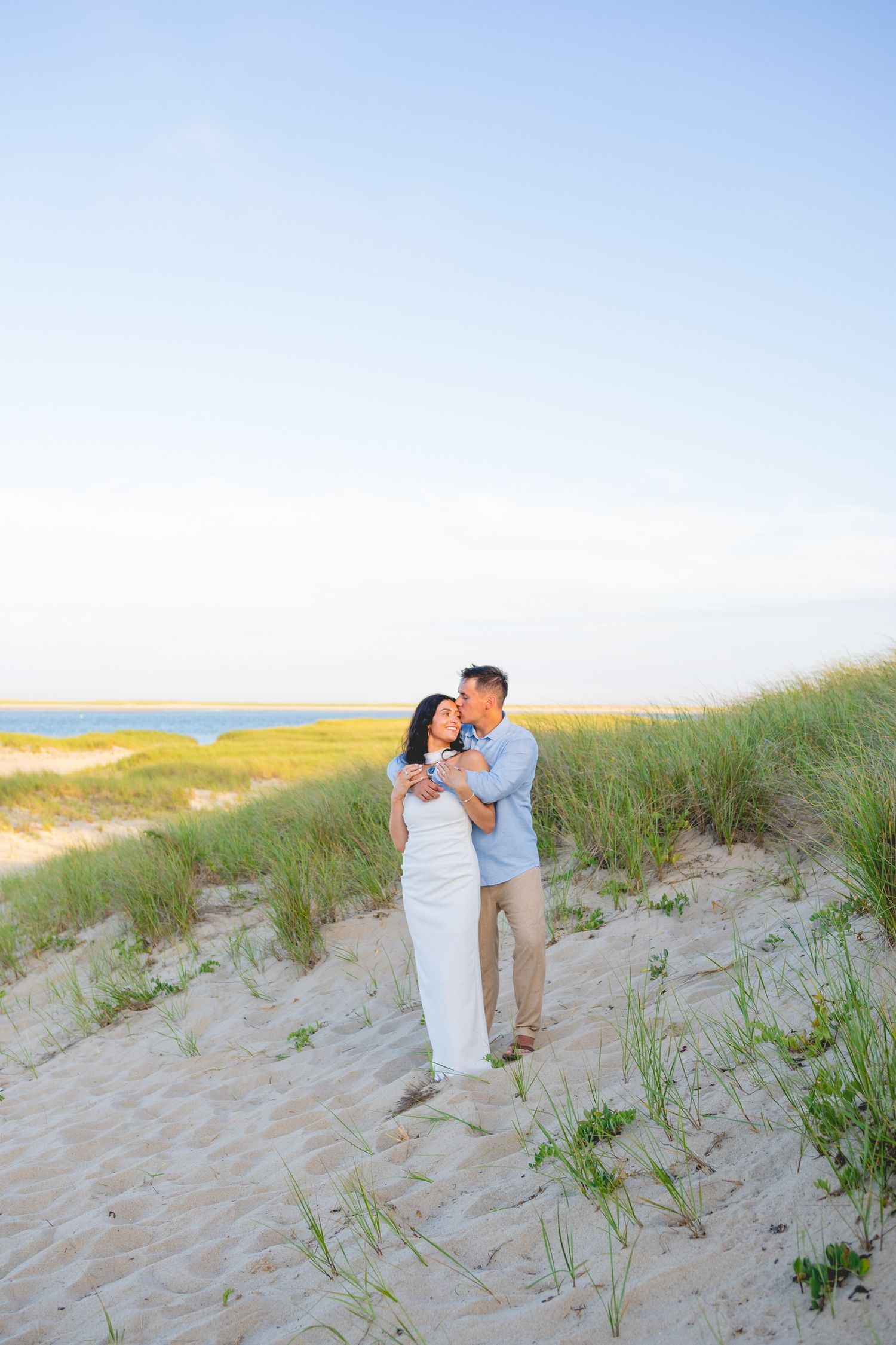 A couple embraces on sandy dunes with beach grass during golden hour in Cape Cod.