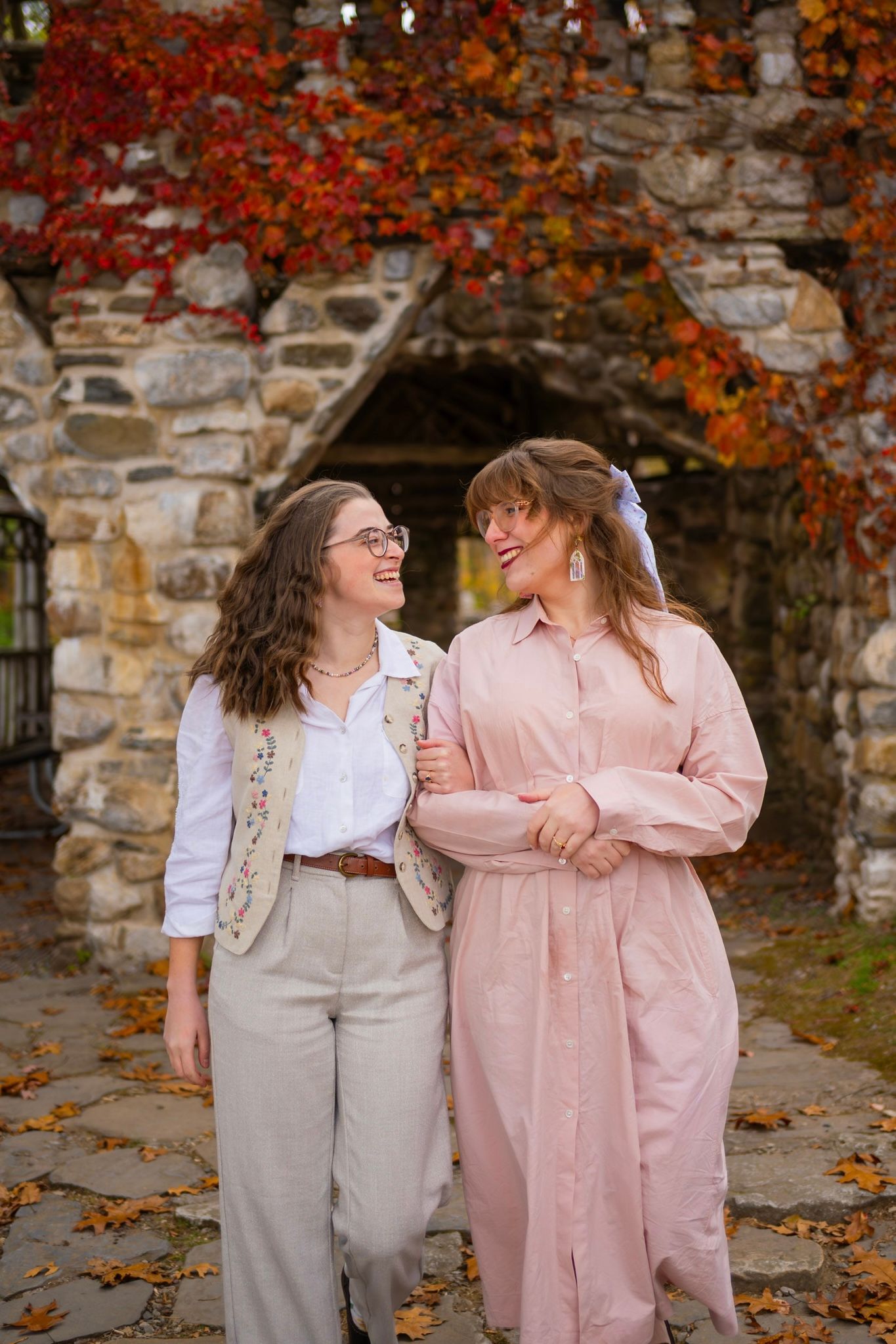 Romantic couple in coordinating pink and beige outfits laughing against a stone wall with fall foliage.