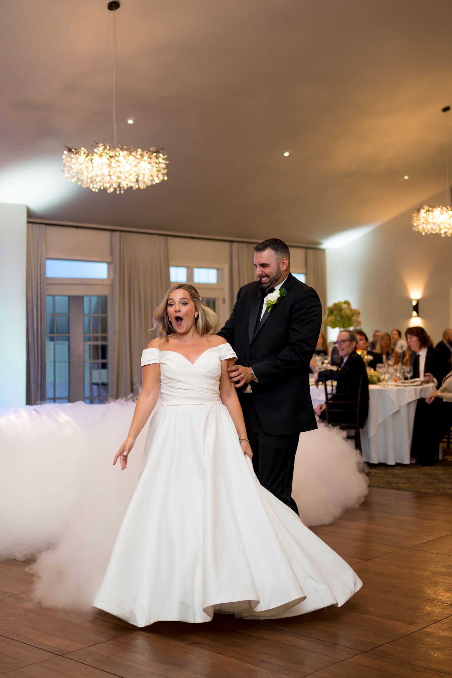Couple in formal wedding attire dancing in an elegant ballroom with crystal chandeliers.